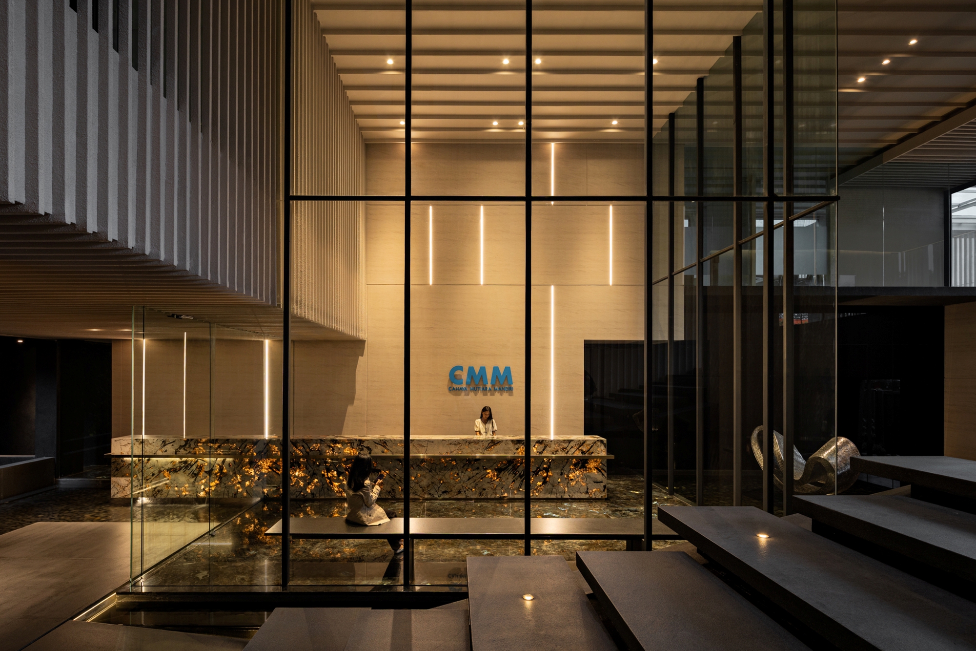 Reception desk with veined stone front seen through a black steel glass grid, with floating stone stairs in the foreground at dusk