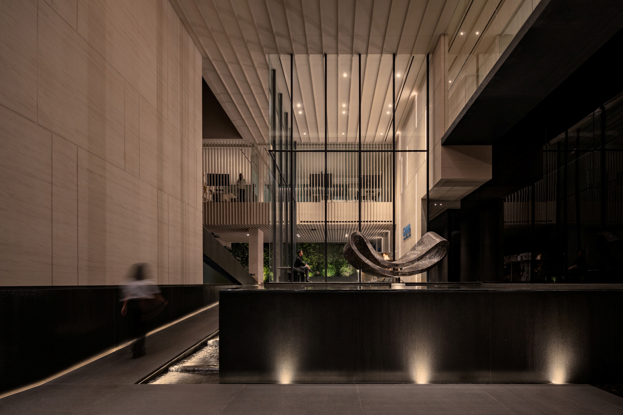 Lobby court with a dark water basin and curved metal sculpture, a blurred figure walking along the lit ramp at night