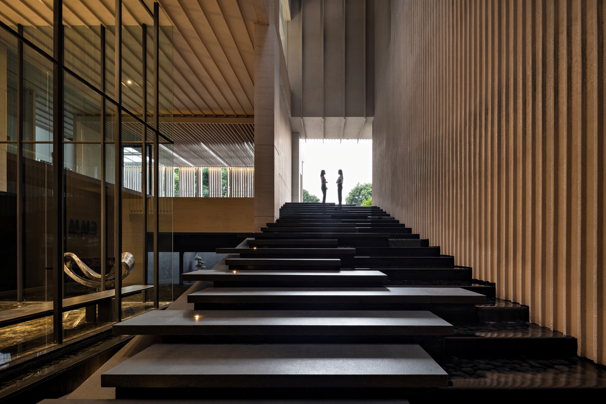Floating black stone stair rising beside a vertically ribbed wall, two figures silhouetted against green daylight at the top