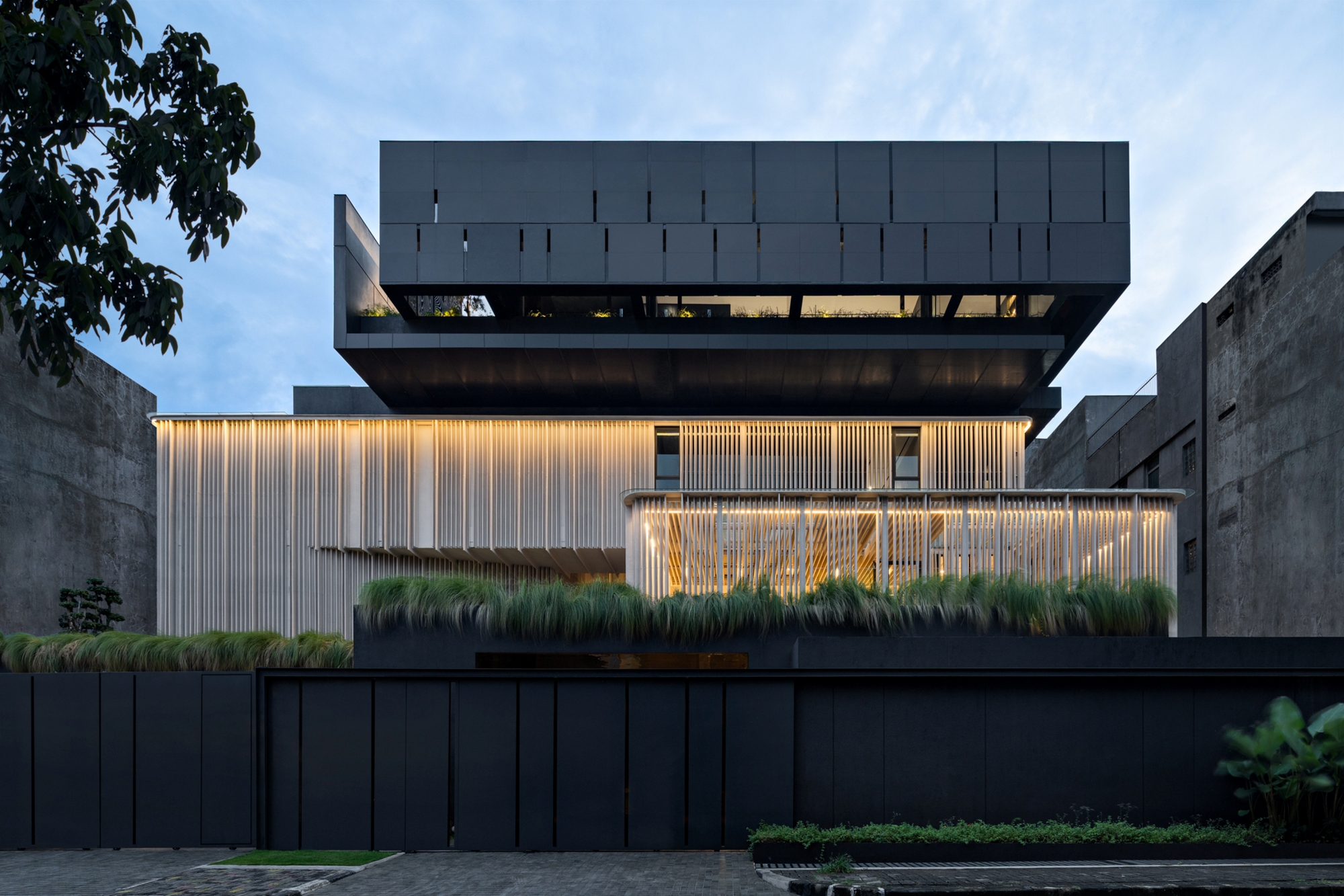 Street facade with a dark cantilevered upper volume above a vertical slatted screen glowing with warm light at dusk, grasses planted along the wall
