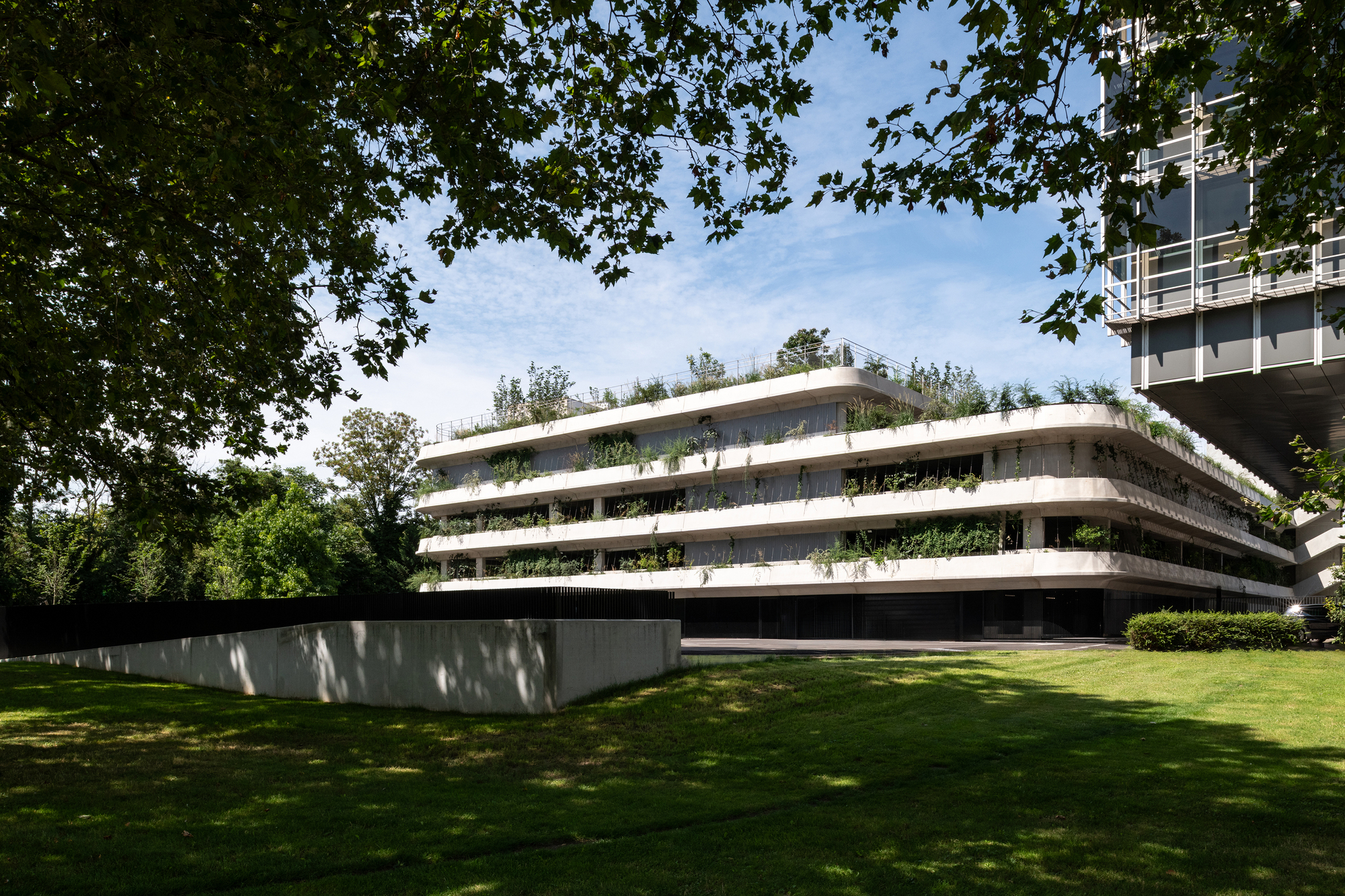 Stepped concrete parking structure with planted terraces on every level, seen across a lawn under tree shade and a bright blue sky