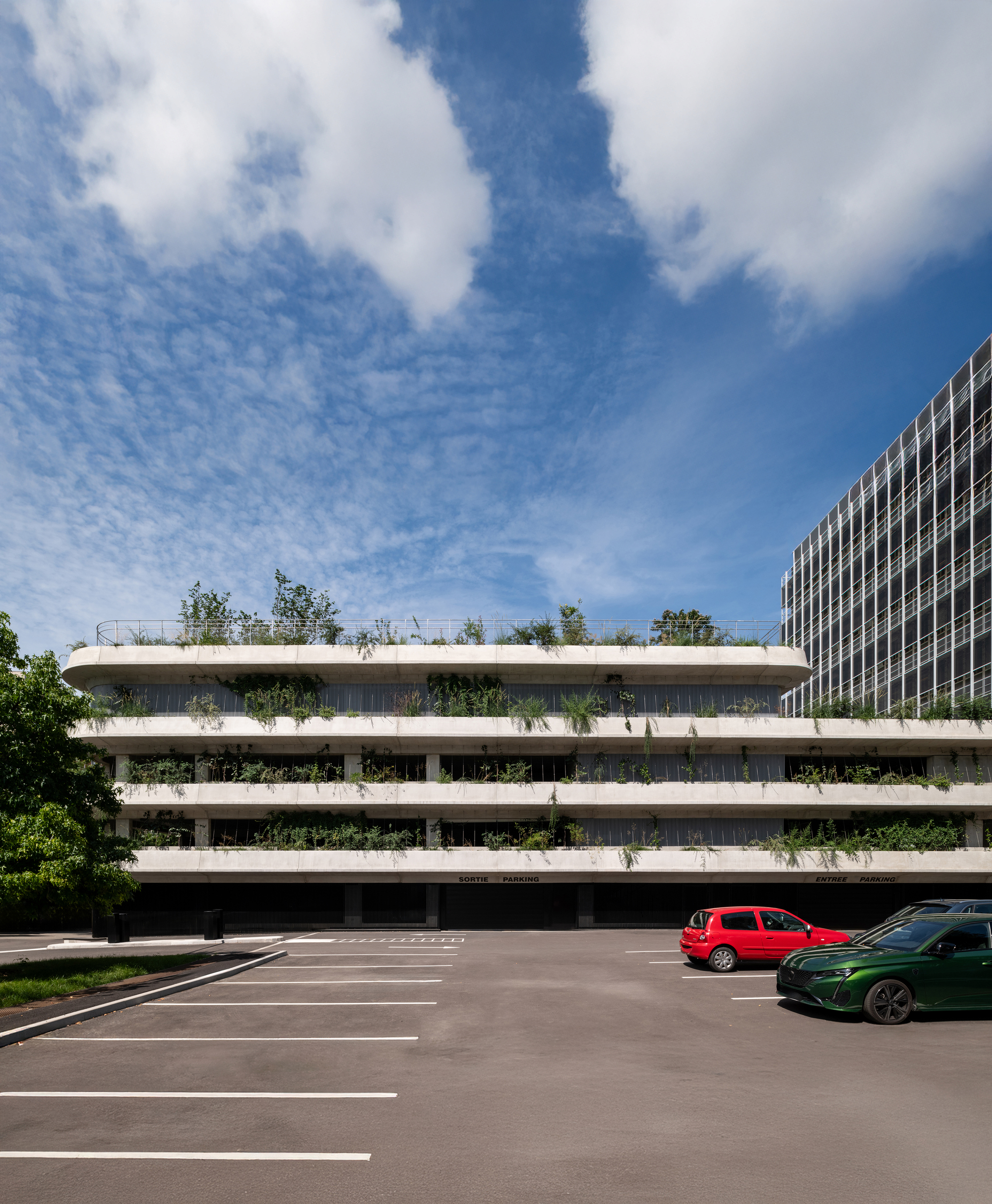 Street-level view of the layered planted parking garage with a red and a green car parked below drifting clouds