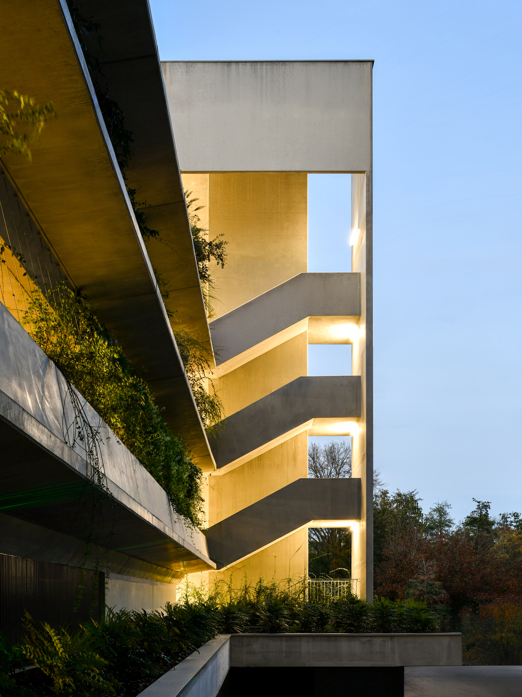 Open concrete stair tower lit from within at dusk, with planted terraces cantilevering in from the left