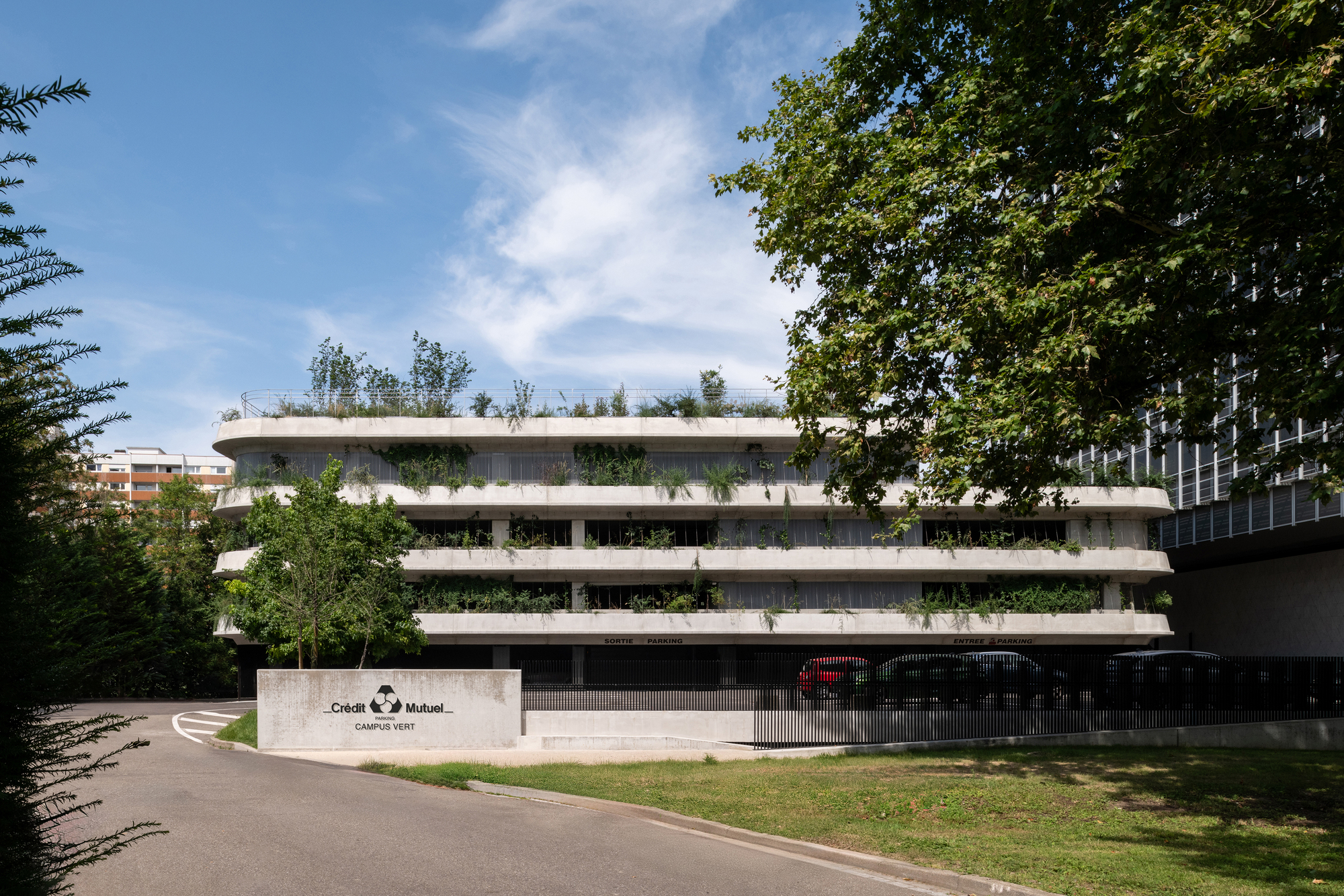 Street-level view of the tiered concrete parking building with planted terraces, parked cars and a sign, framed by tree branches