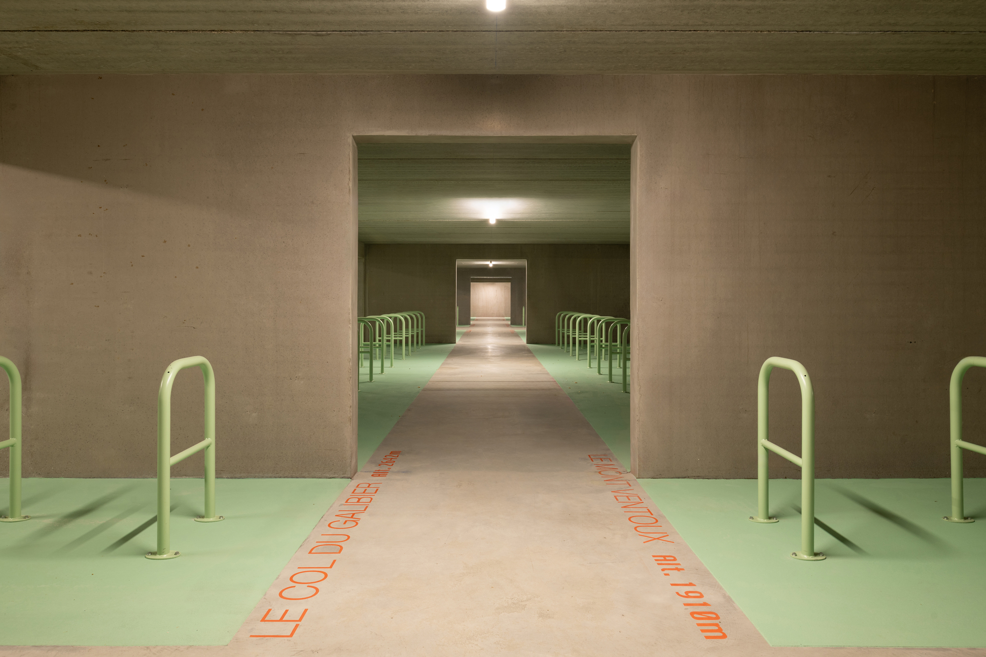 Concrete bicycle parking hall with pale green floor, green steel racks and orange painted lettering leading down the central path to a doorway