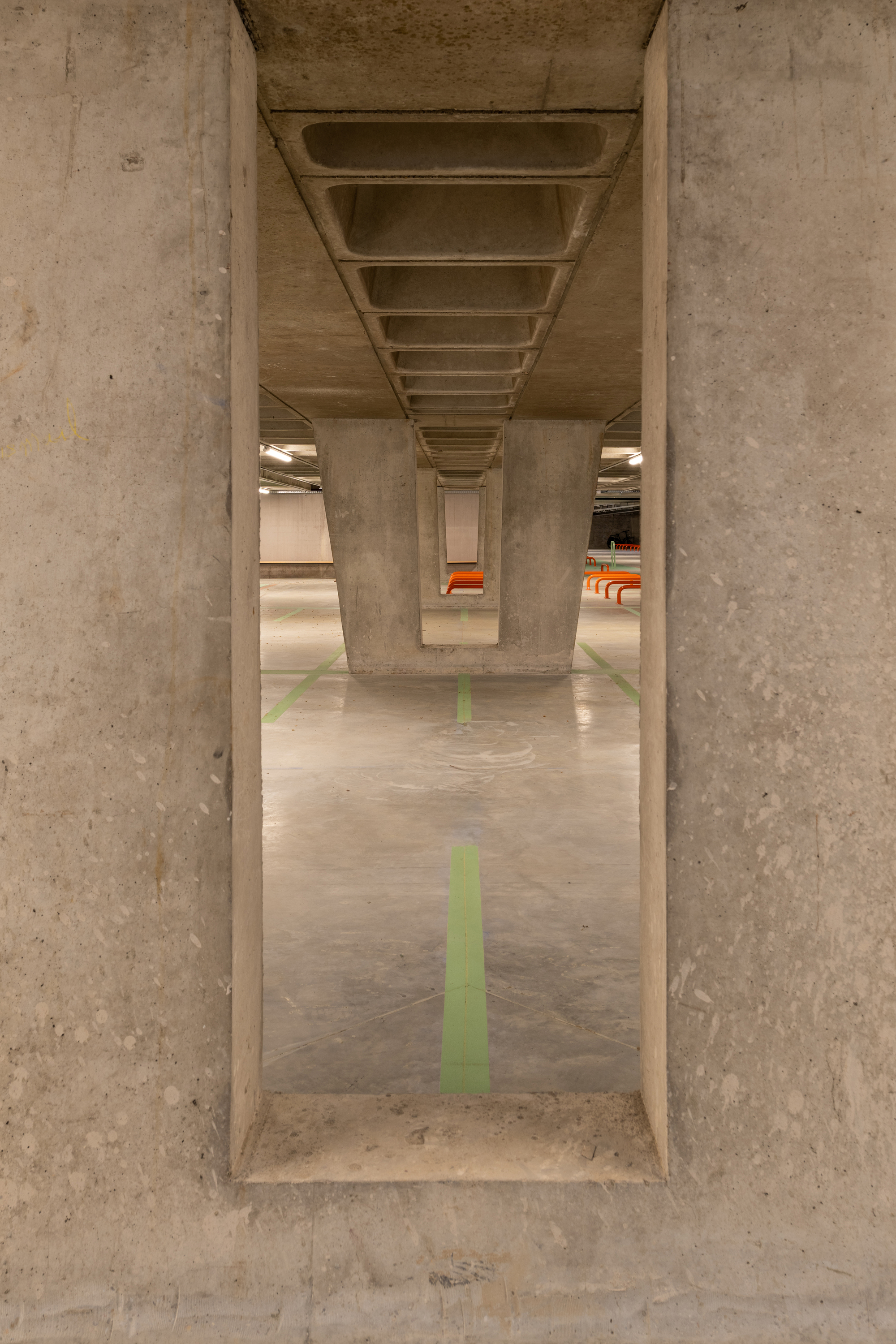 Framed view through a deep concrete wall opening to a ribbed slab, tapered columns and green floor lines in dim artificial light