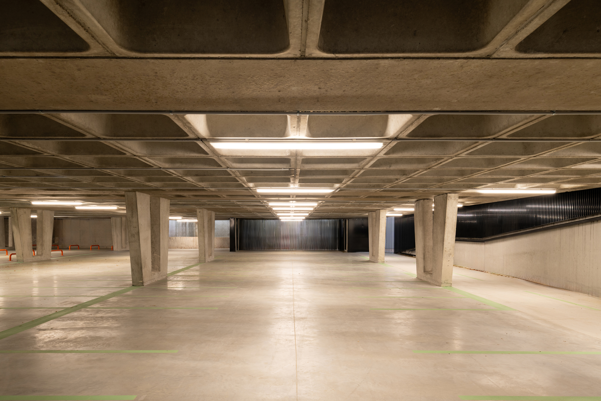 Underground parking level with a waffle concrete ceiling, tapered columns and strip lights above a pale floor marked with green lines