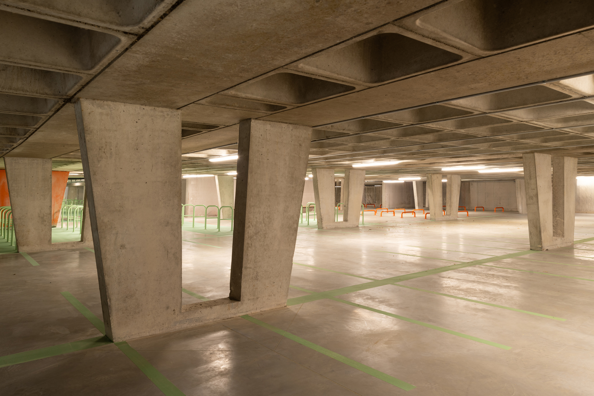 Underground bike parking hall with tapered concrete columns, coffered slab ceiling and green floor markings under warm lamp light