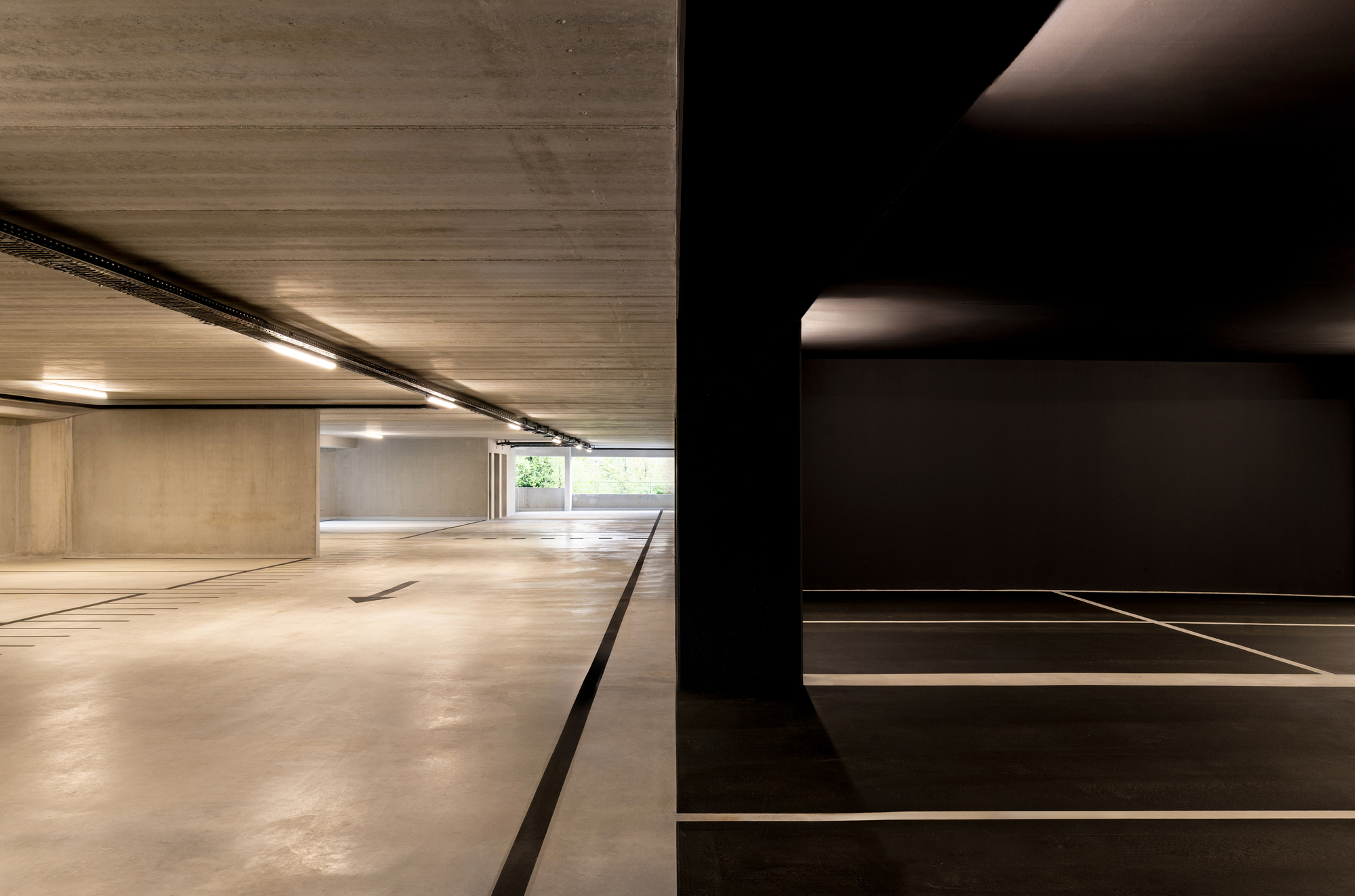 Garage level where a black-painted bay with white stall lines meets a raw concrete aisle, with green foliage glimpsed at the far end