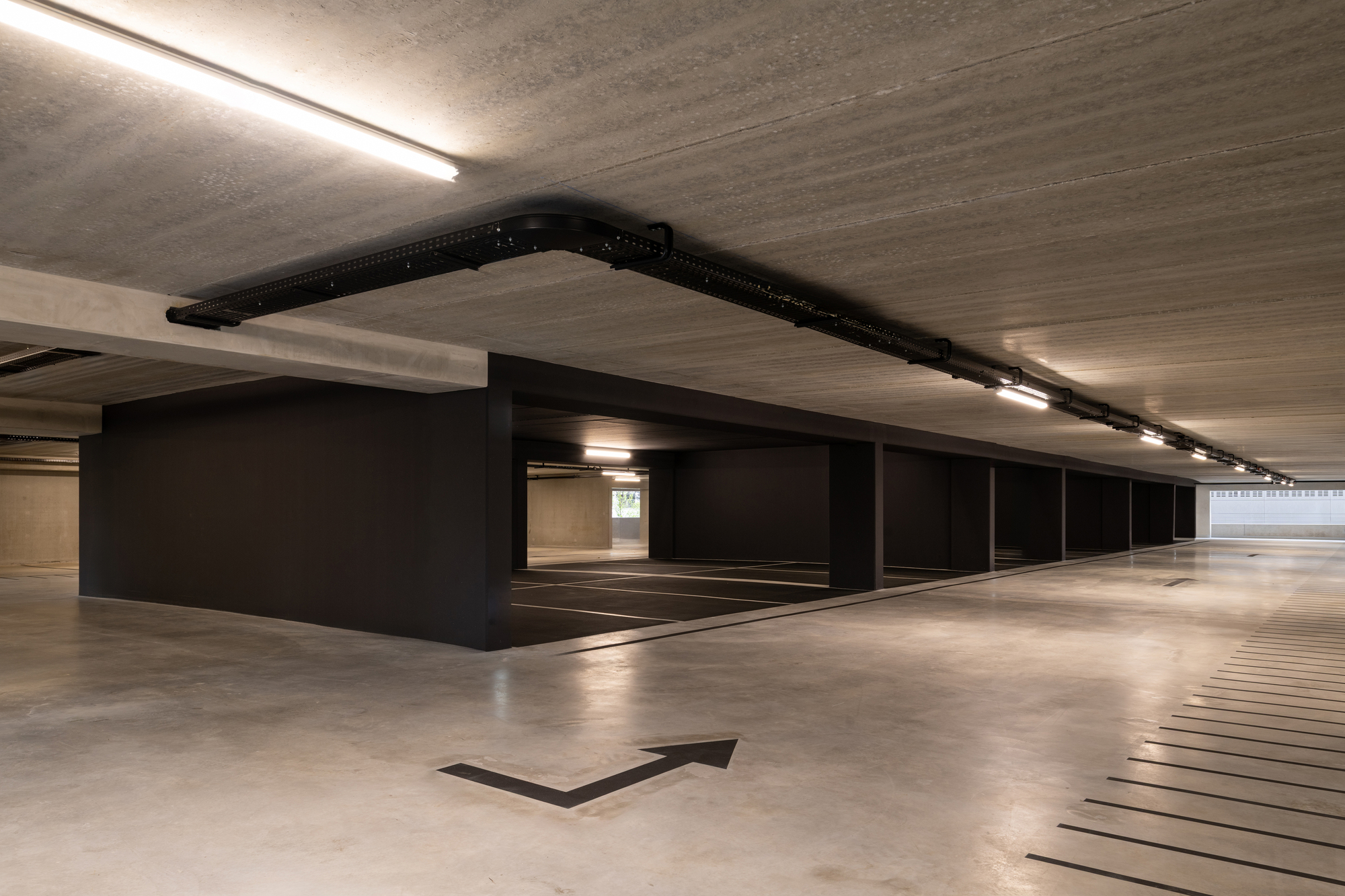 Parking floor with dark-clad bays, a black cable tray along the board-marked concrete ceiling and a painted turn arrow on the floor