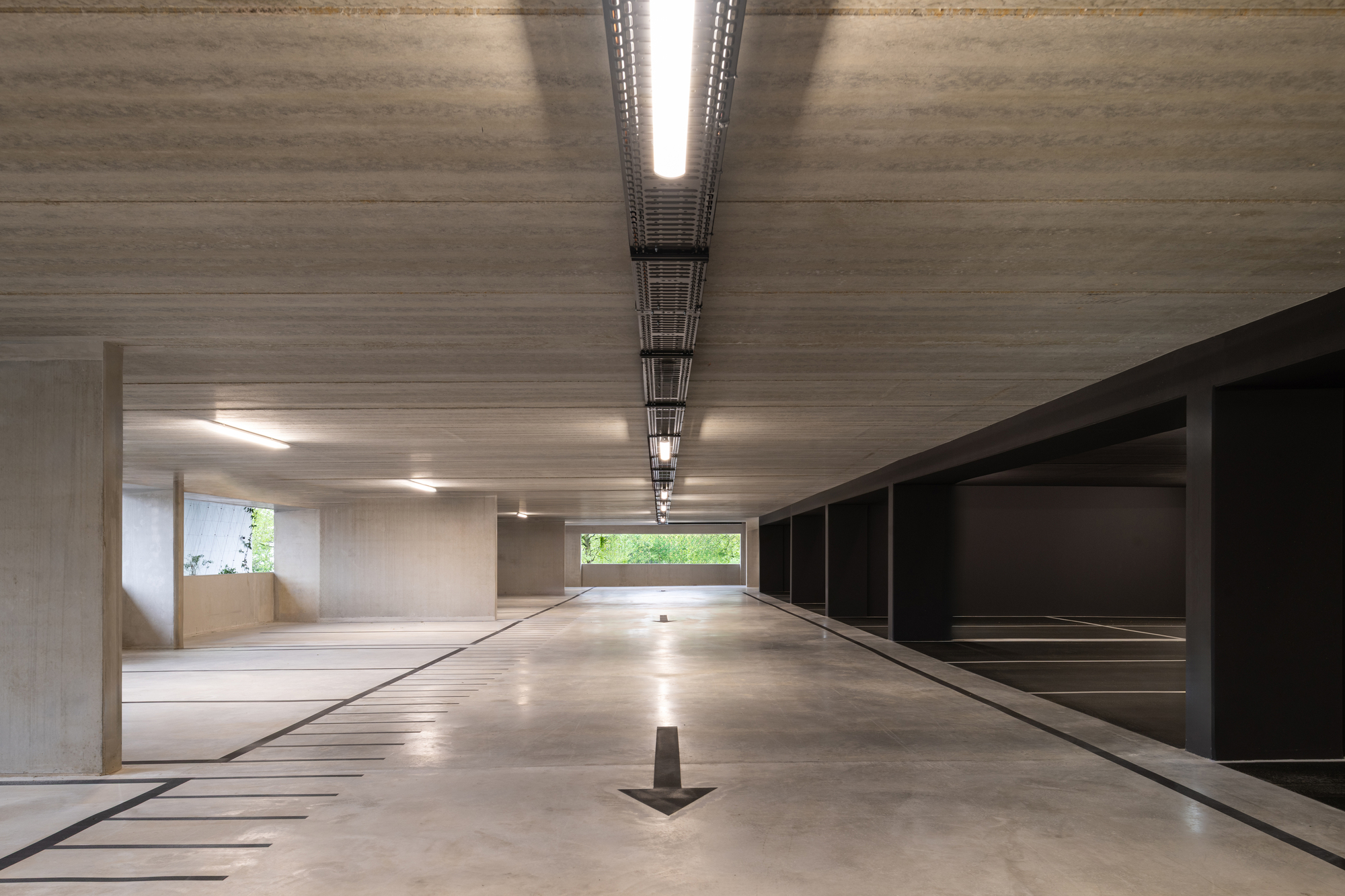 Central drive aisle in the garage with a single light strip overhead and a painted arrow, daylight and greenery at the far opening