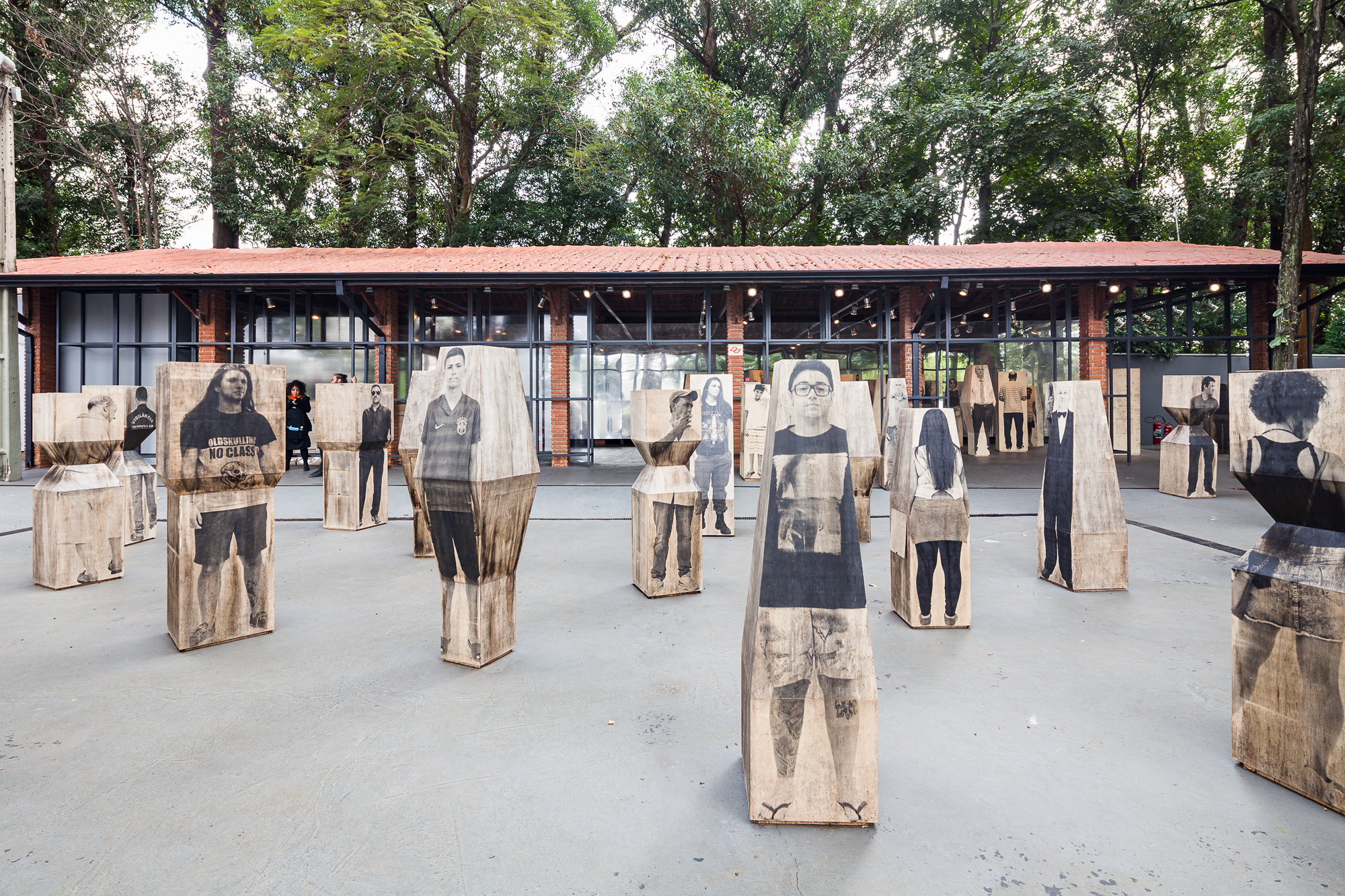 Wooden blocks printed with life-size standing portraits spread across a paved plaza in front of a brick pavilion with a red tile roof