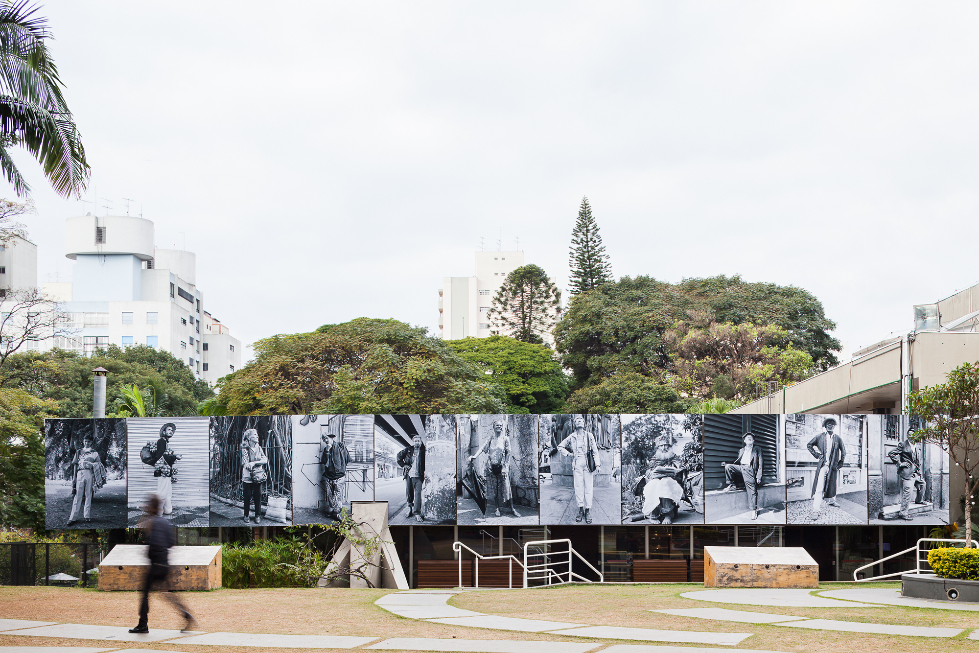 Long band of black and white street portraits mounted above a low pavilion, with a blurred pedestrian passing on the lawn