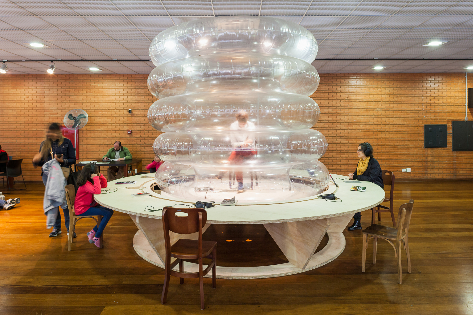 Circular plywood table around a stack of clear inflatable rings, with visitors reading under a brick wall