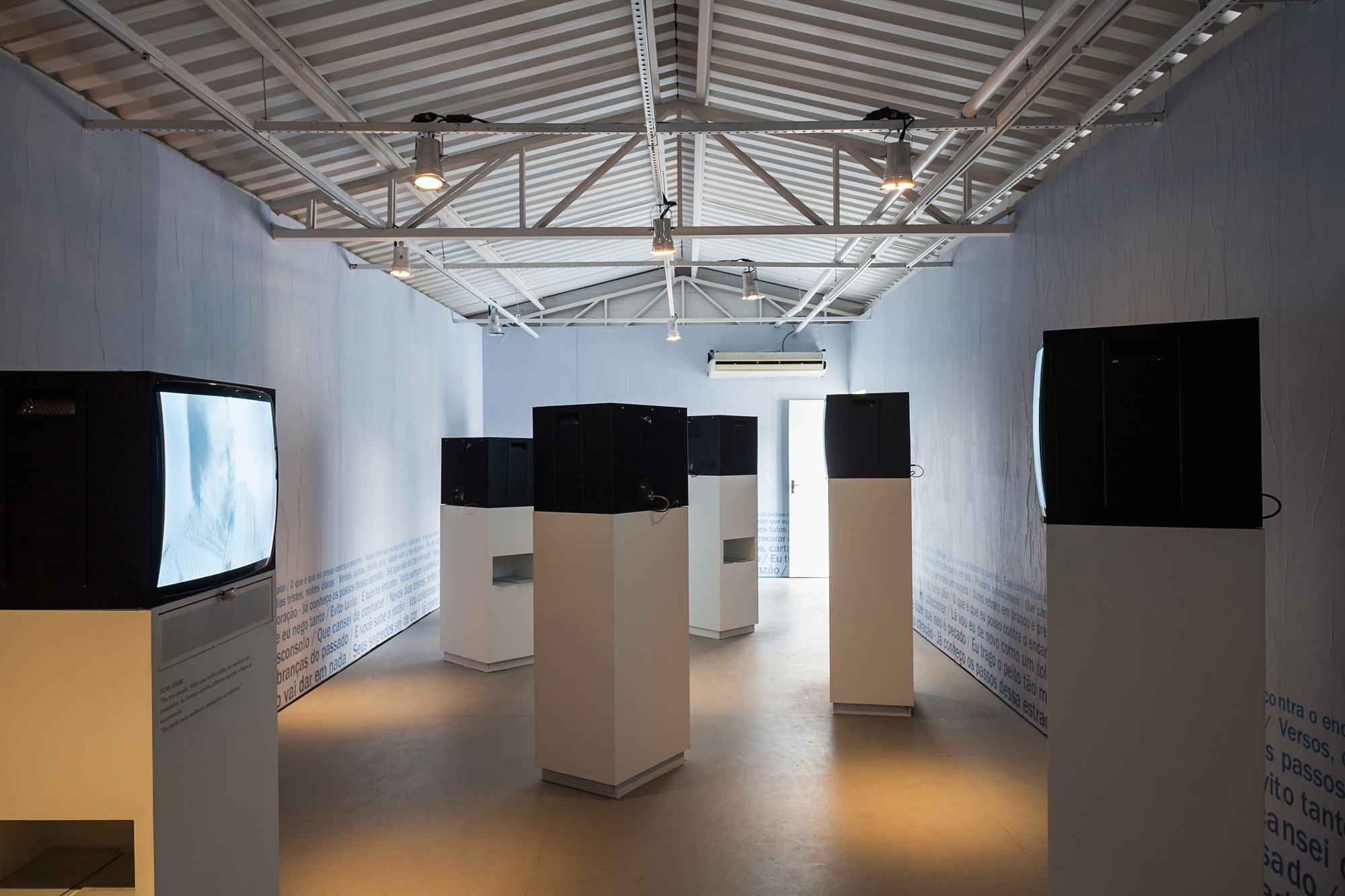 Exhibition hall with old television monitors on white plinths under exposed steel roof trusses and text-covered walls