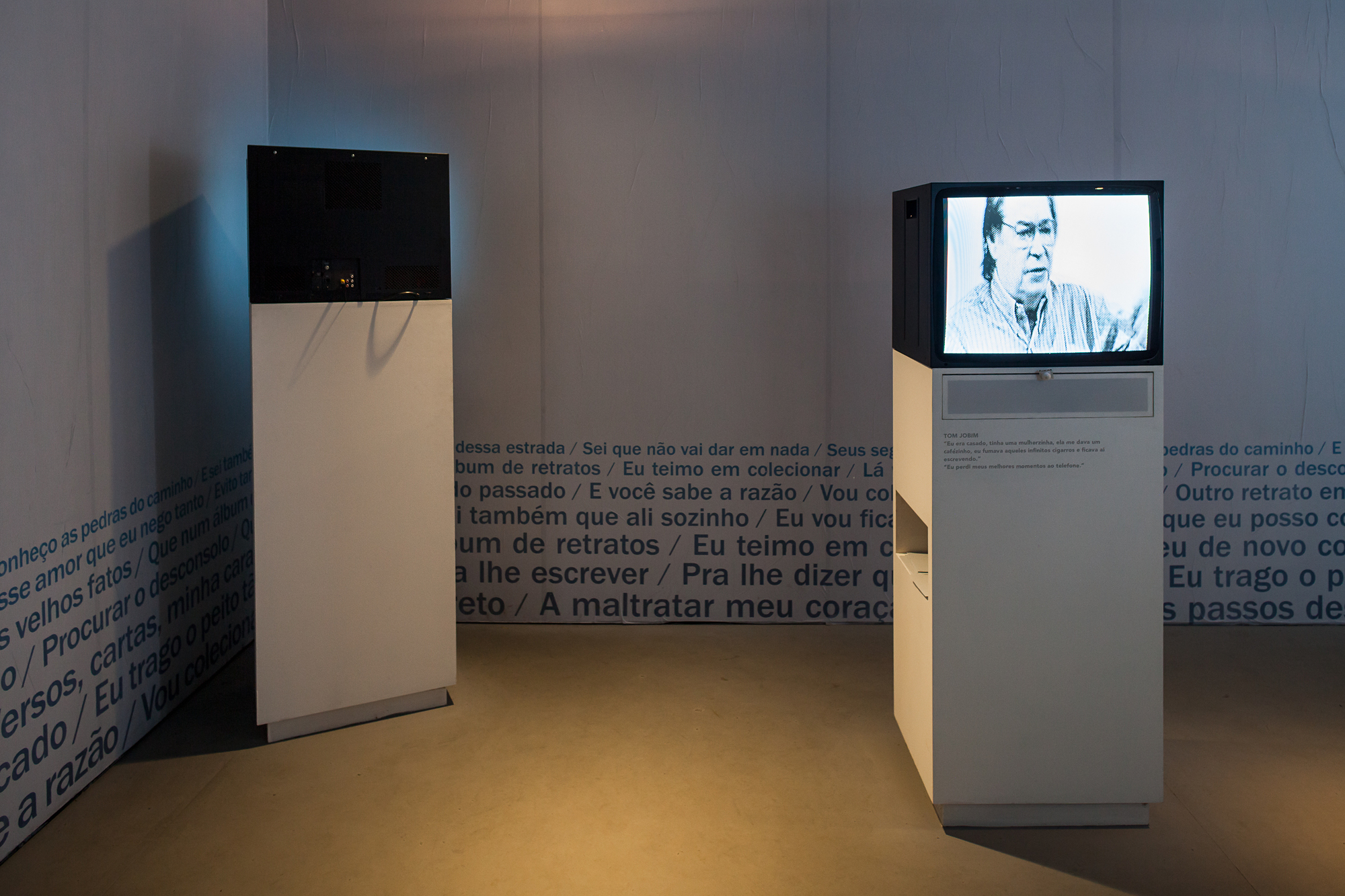 Two white pedestals with video monitors, one showing a black-and-white interview, before walls printed with lines of song lyrics in dim light