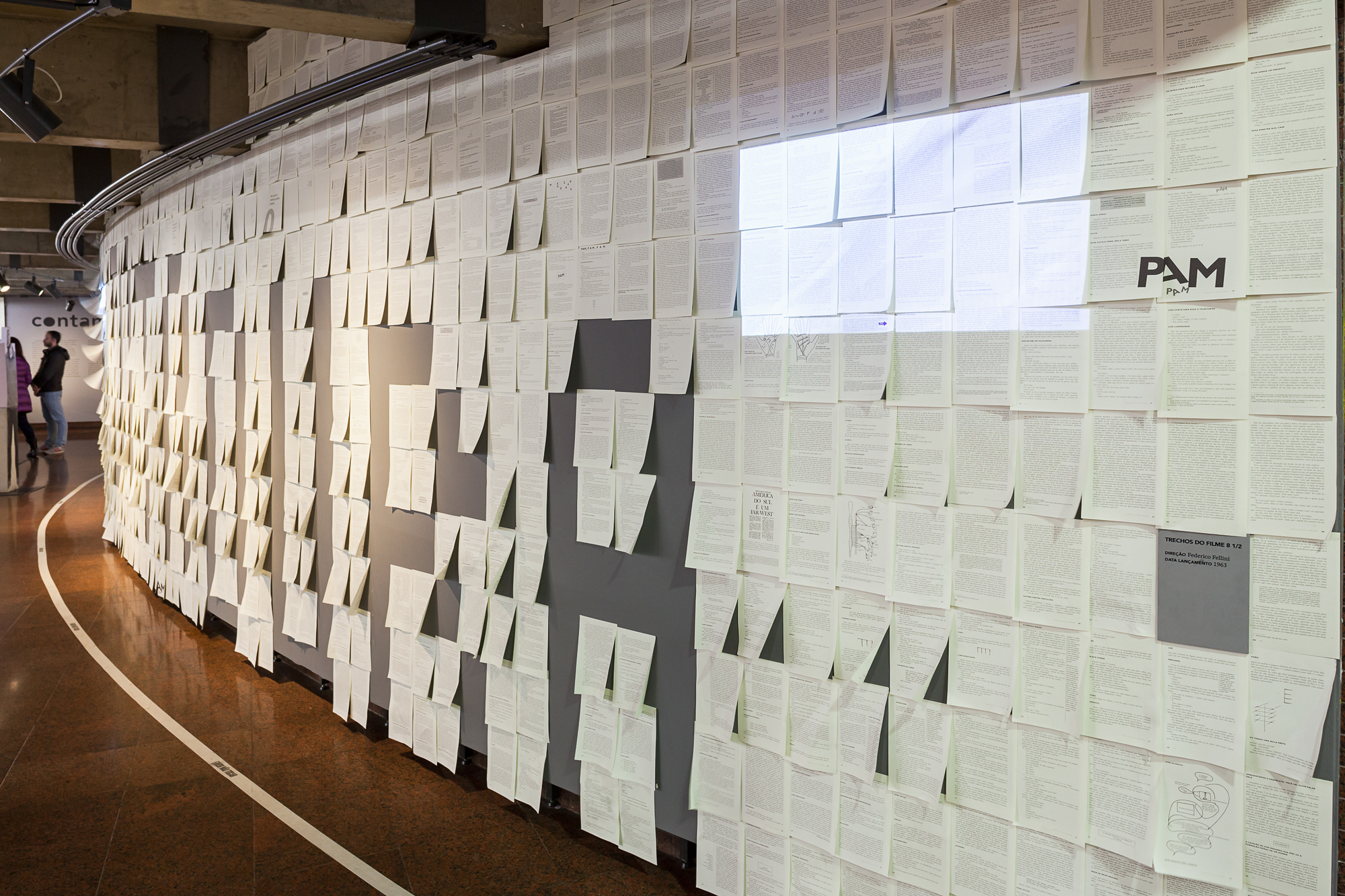 Curved wall covered in rows of pinned typed pages, with a projected rectangle of light across the upper sheets