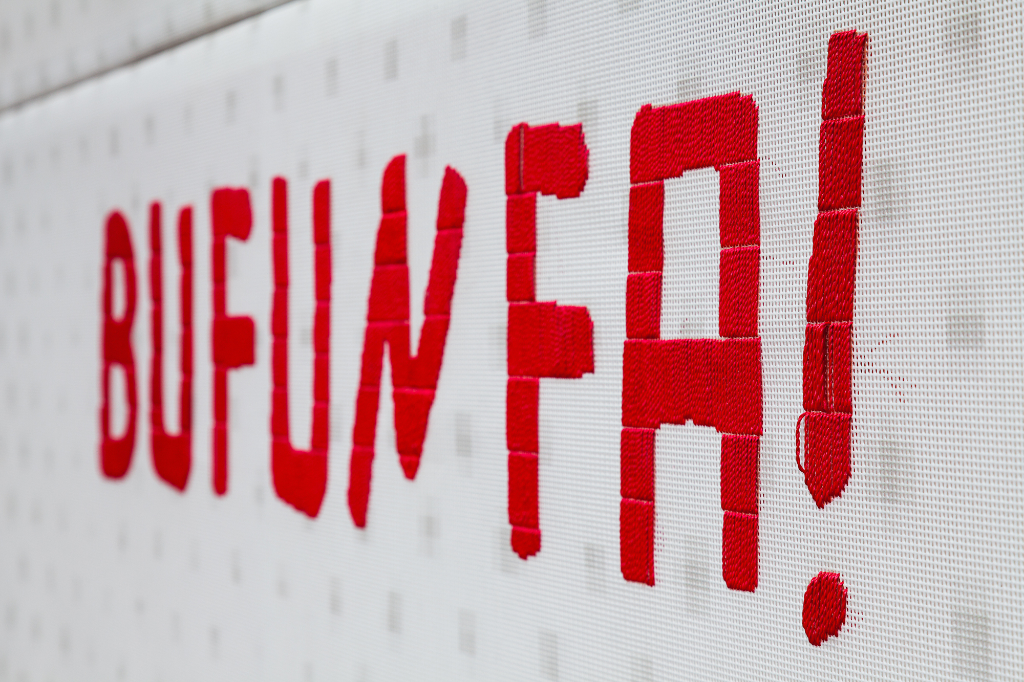 Close-up of red embroidered capital letters spelling a word with an exclamation mark on white perforated fabric, seen at a sharp angle