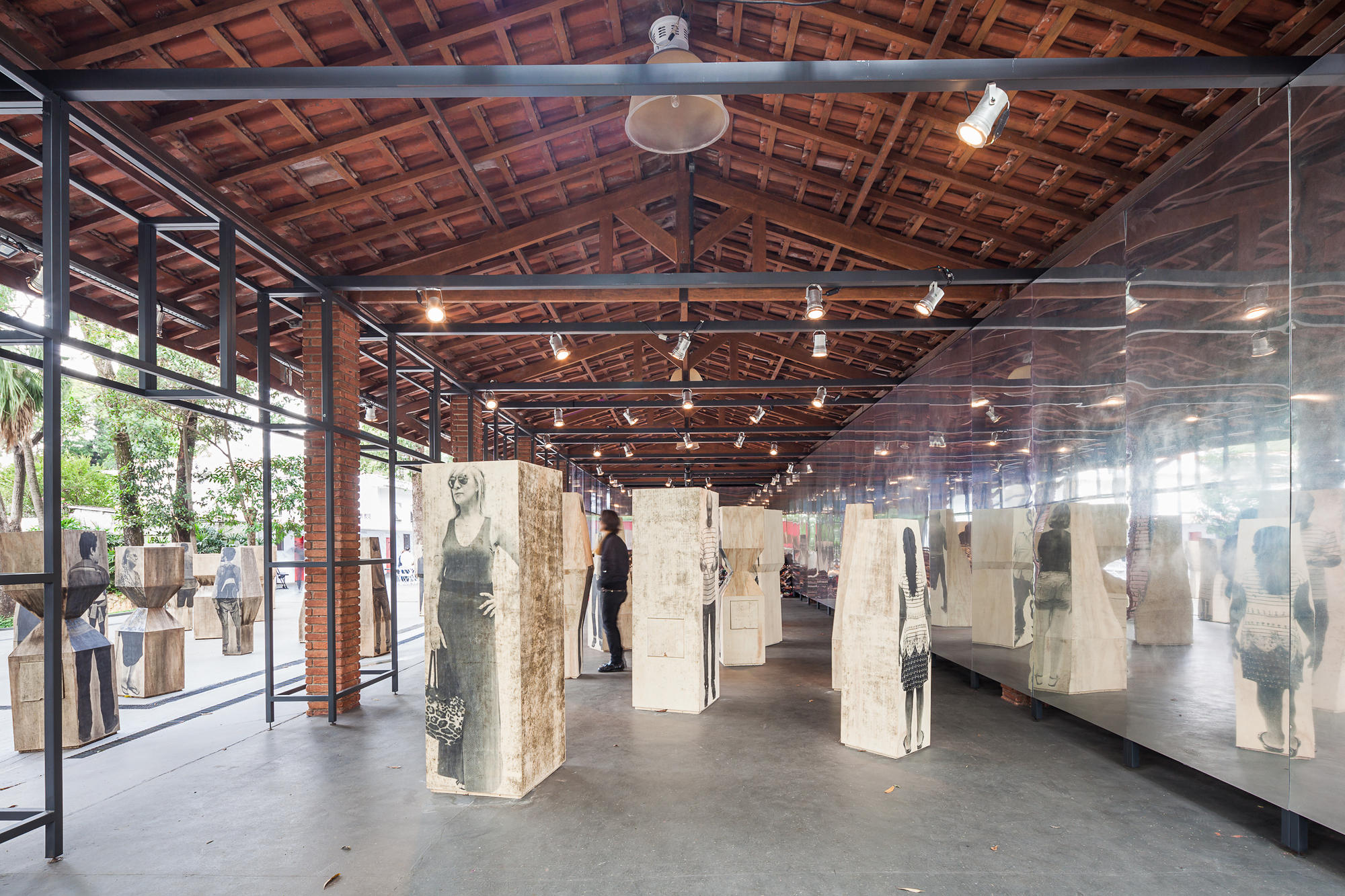 Steel-framed pavilion under a timber and clay tile roof, with printed wooden blocks standing in rows beside a mirrored wall