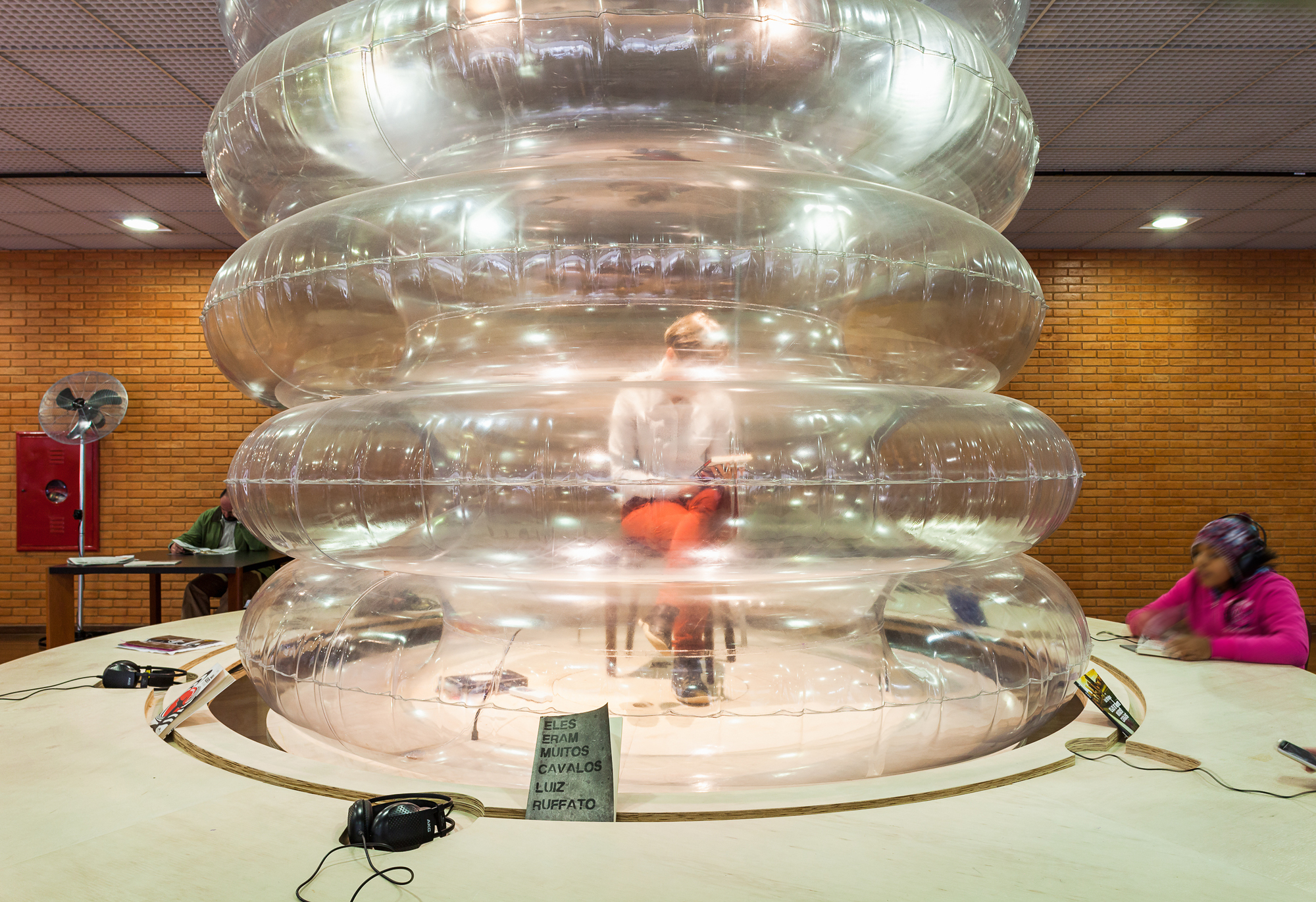Stacked clear inflatable rings on a round wooden table, with a seated reader visible inside and others nearby against a brick wall