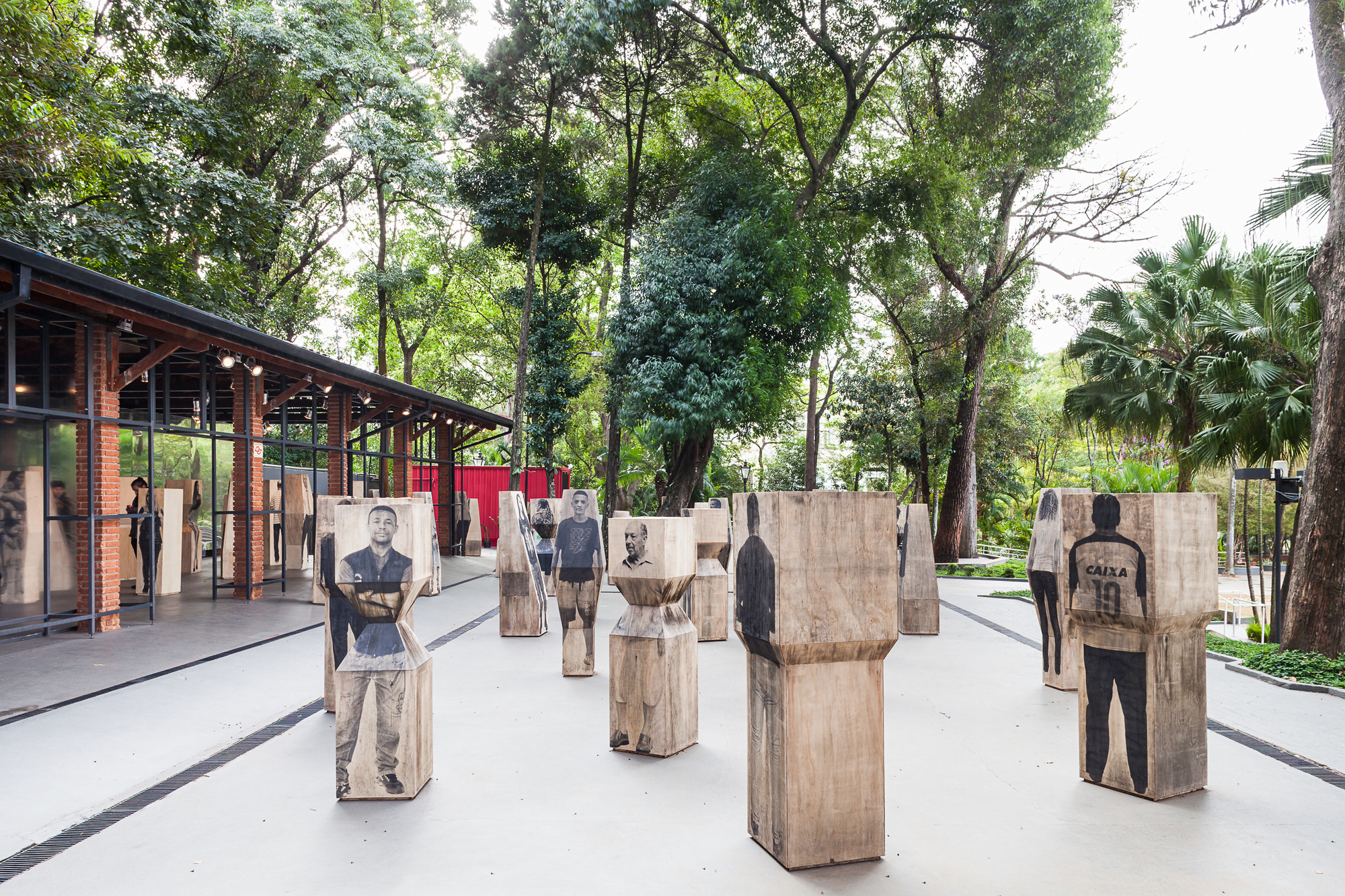 Wooden pedestals printed with life-size portraits stand on a paved plaza beside a glazed brick pavilion under tall trees