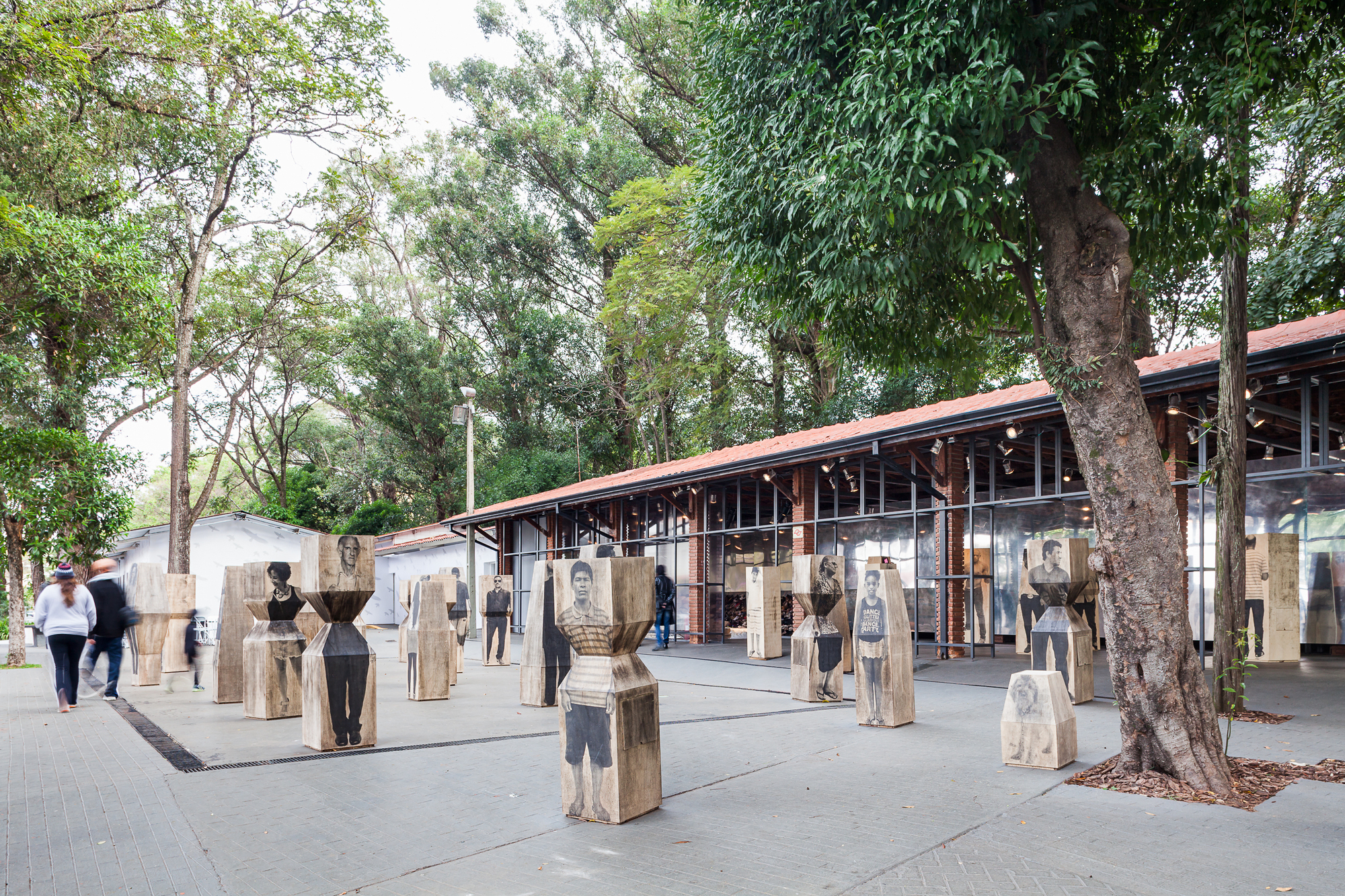 Portrait-printed timber blocks scattered across a paved square in front of a glazed pavilion, with visitors walking past under a large tree