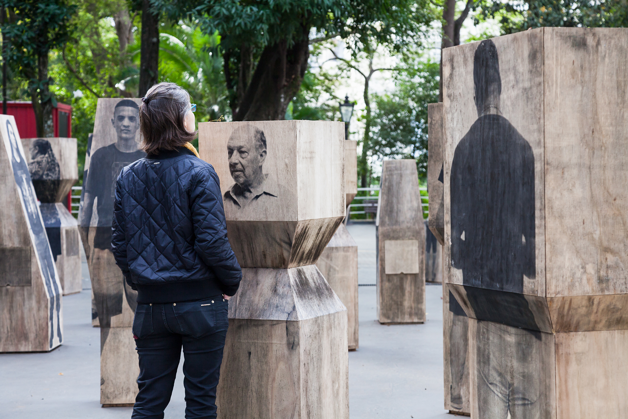 Visitor in a quilted jacket studies an elderly man's face printed on a hourglass-shaped timber block in overcast light