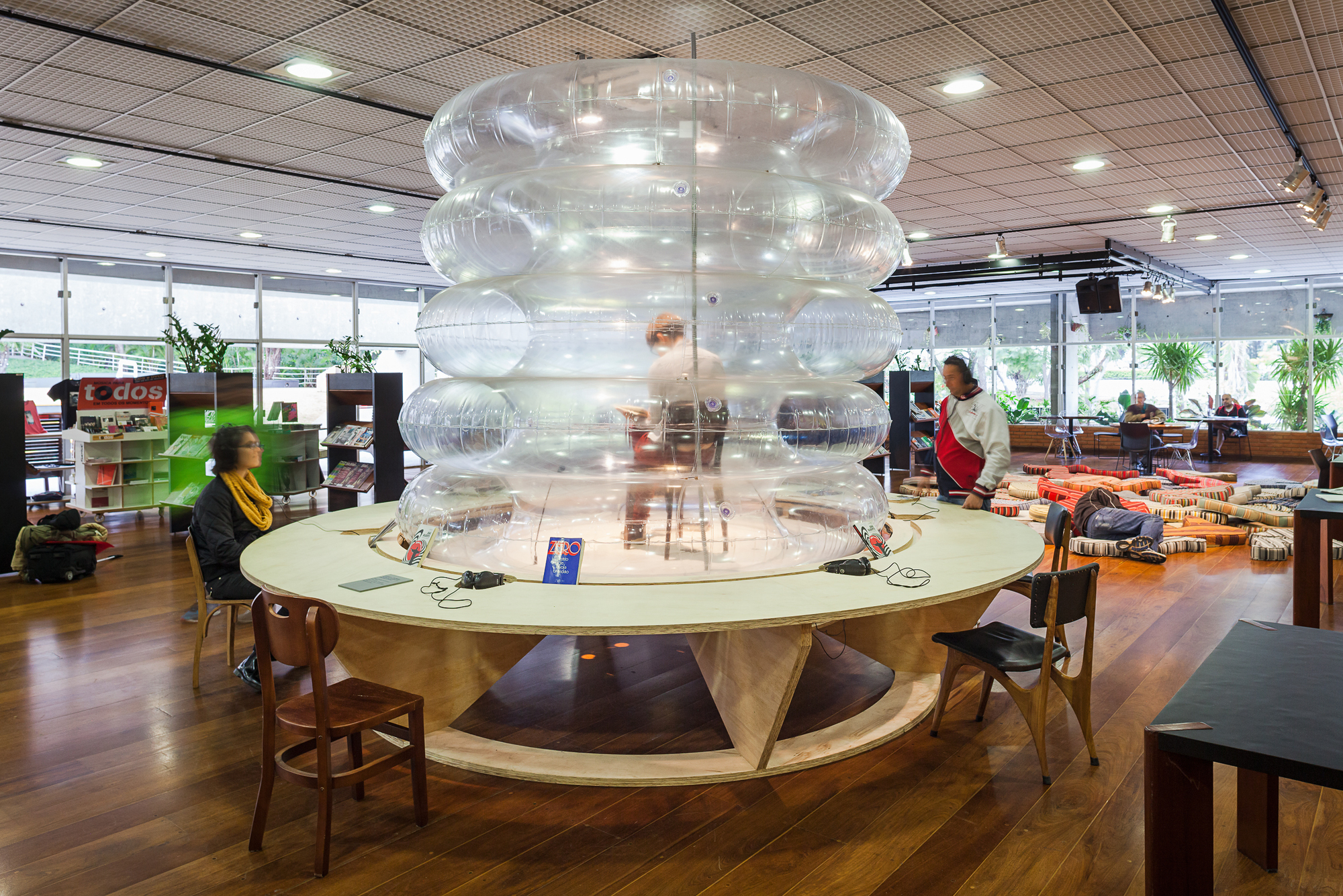 Inflatable clear plastic tower rising from a round plywood table in a library reading room with wood floors and floor cushions