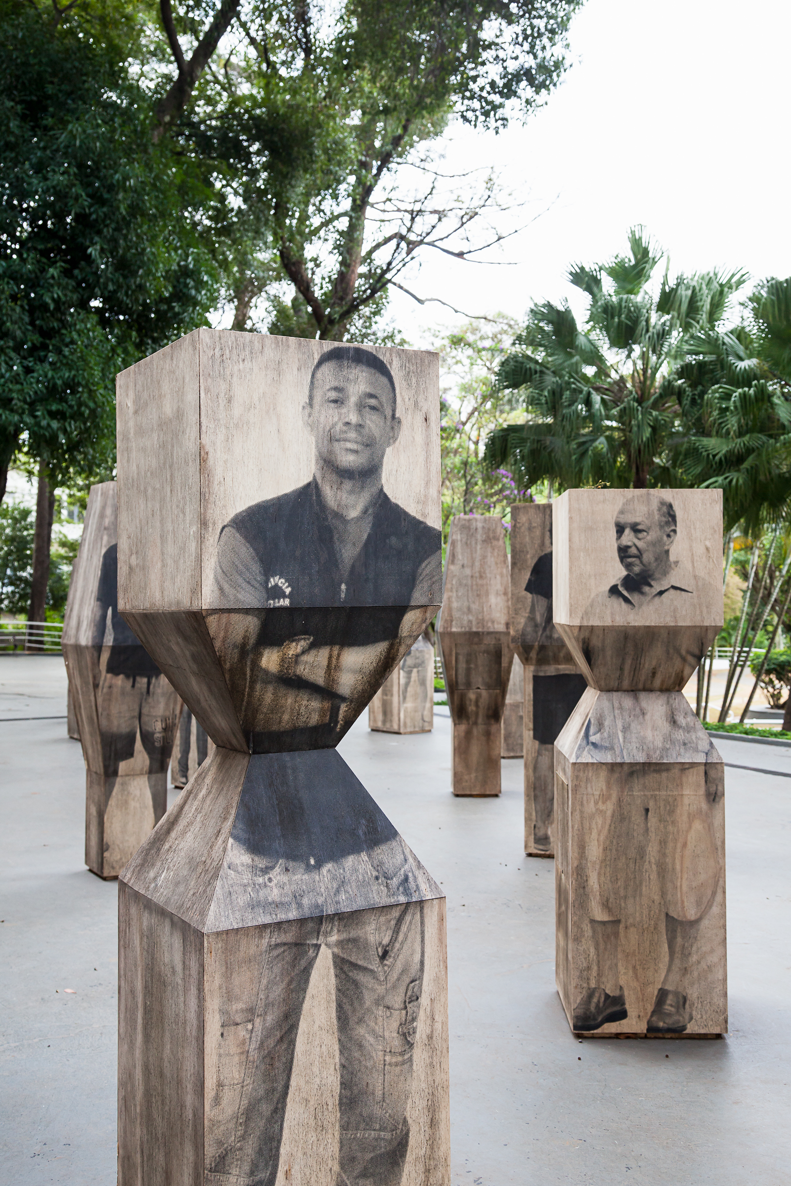 Close view of two timber pedestals printed with full-length portraits of men, with palms and trees behind in soft daylight