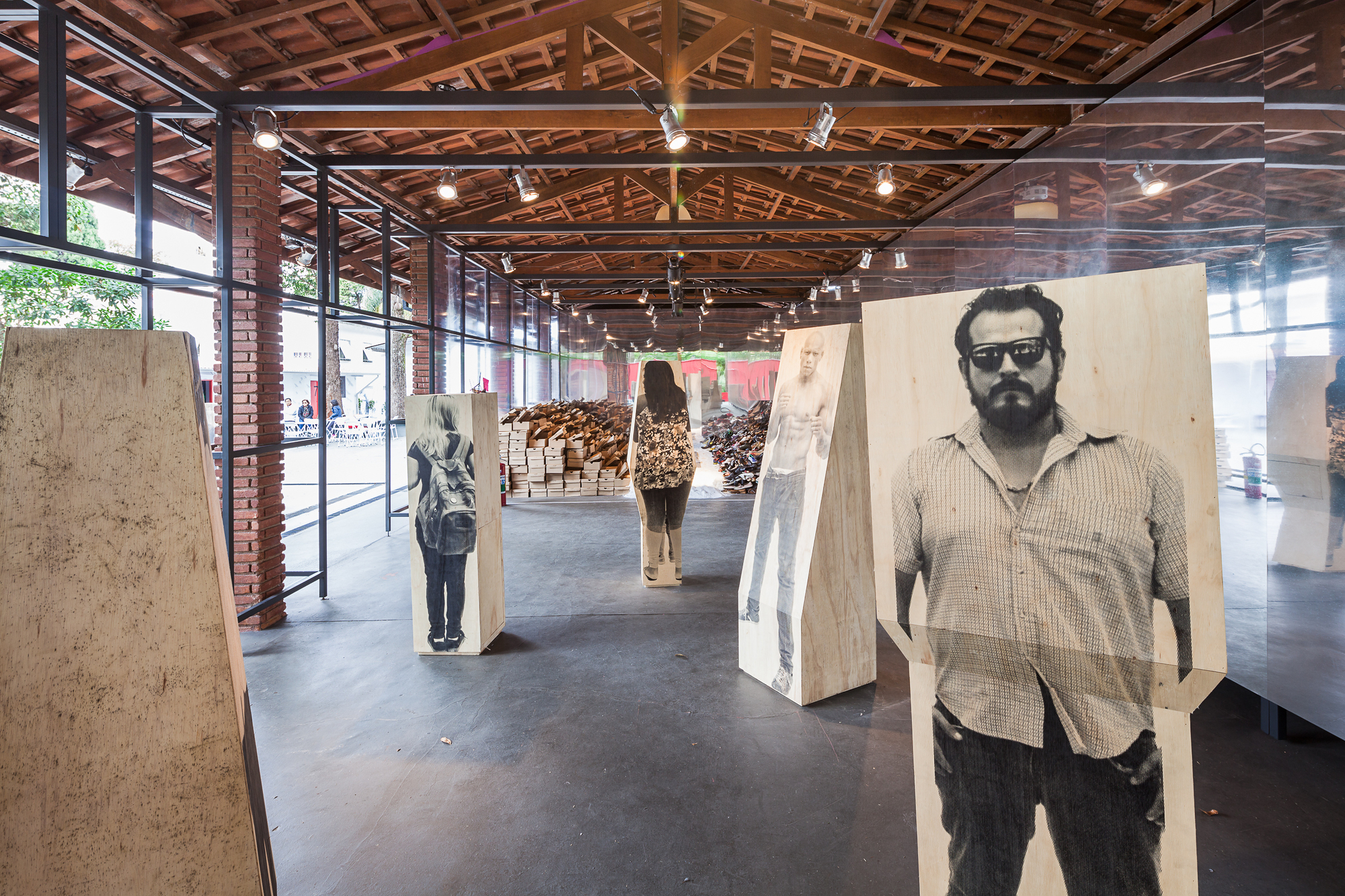 Exhibition hall with plywood-mounted portrait photographs standing in a row beneath exposed timber roof trusses and a visitor walking toward stacked wood