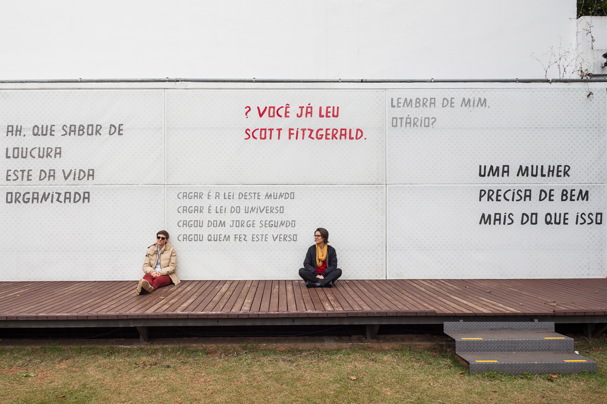 Two seated people on a timber deck in front of a white wall printed with Portuguese text panels