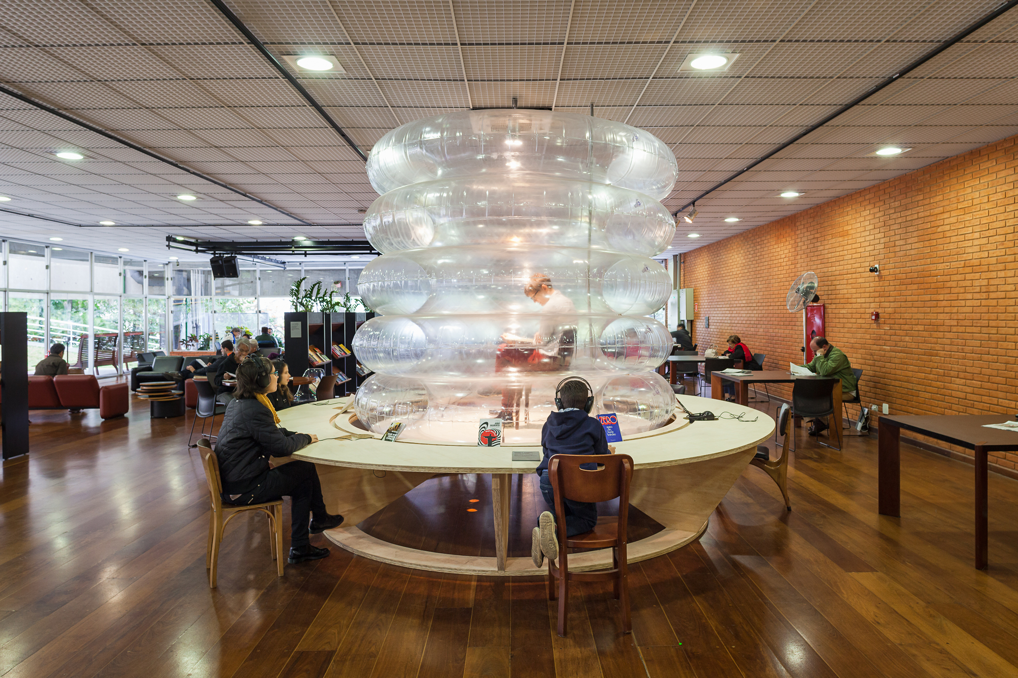 Library reading room with a stacked clear inflatable column rising from a round plywood table, a child in headphones seated at its edge