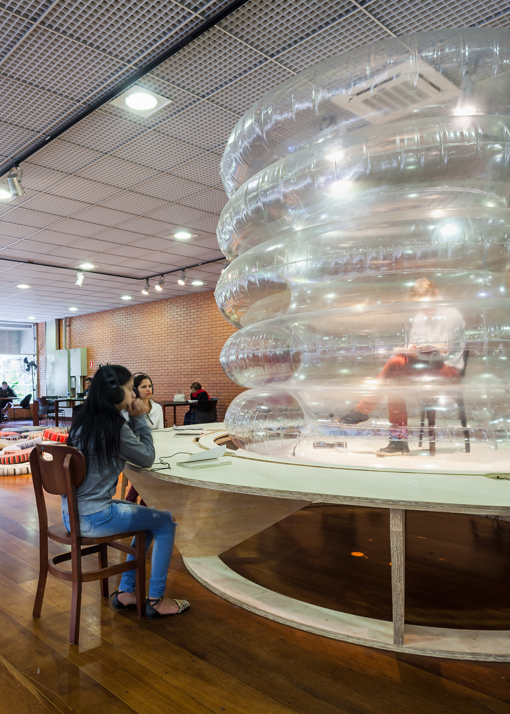 Close view of the translucent inflatable rings above a round table, with a young woman in headphones seated beside a brick wall