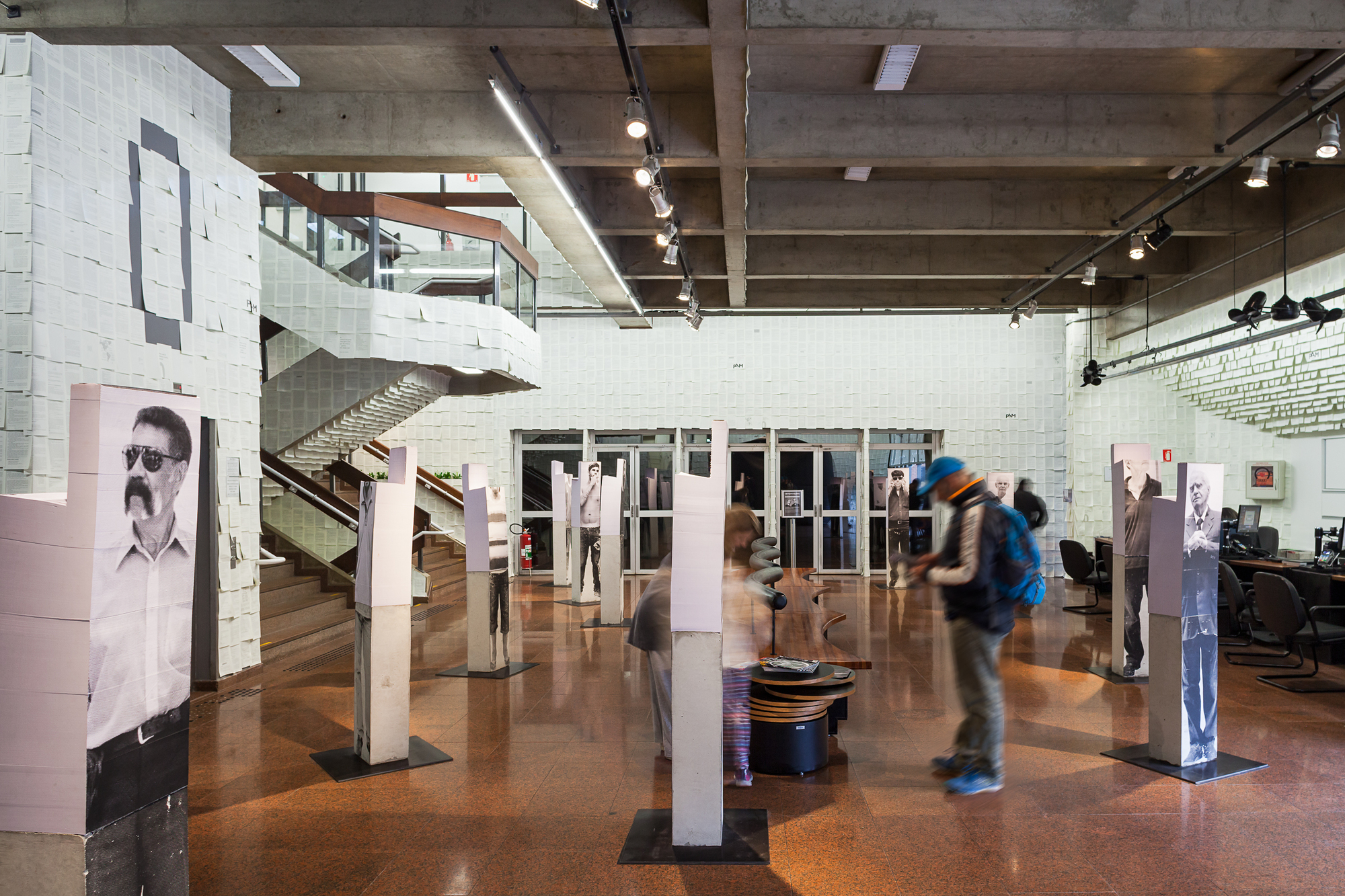 Gallery hall with tall photo-printed columns, a paper-clad staircase and a visitor in a blue cap passing in motion blur