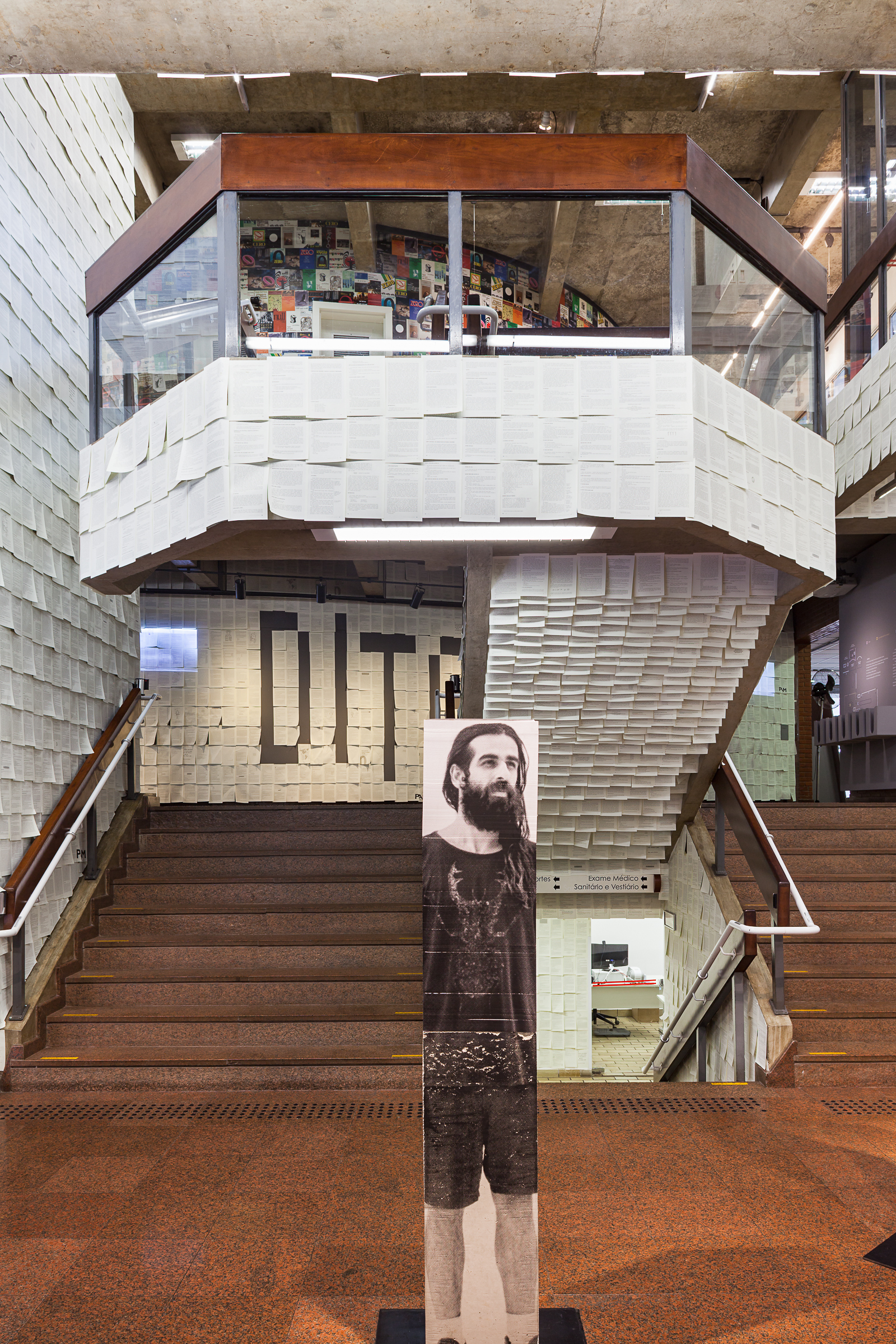 Paper-clad stair landing with a glazed timber-framed balcony and a portrait column of a bearded man in the foreground