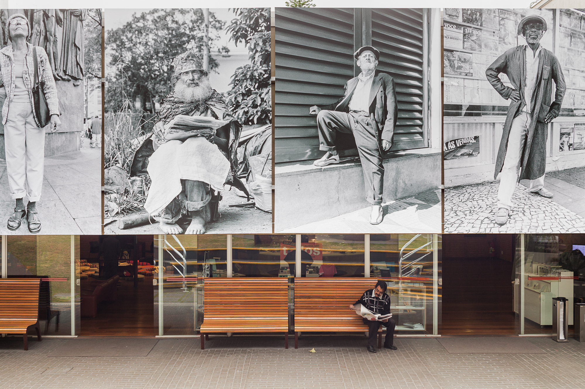 Large black-and-white street portraits above a glazed ground floor where a man reads a newspaper on a wooden bench