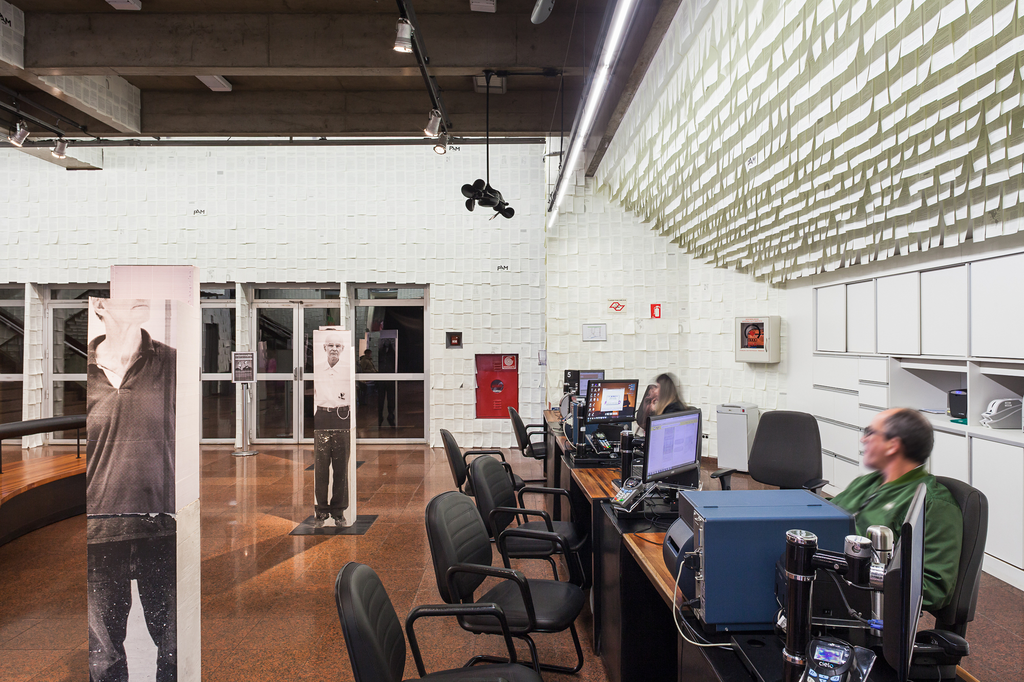 Reception desk with computers and seated staff beside a wall of layered paper sheets and portrait columns under track lights