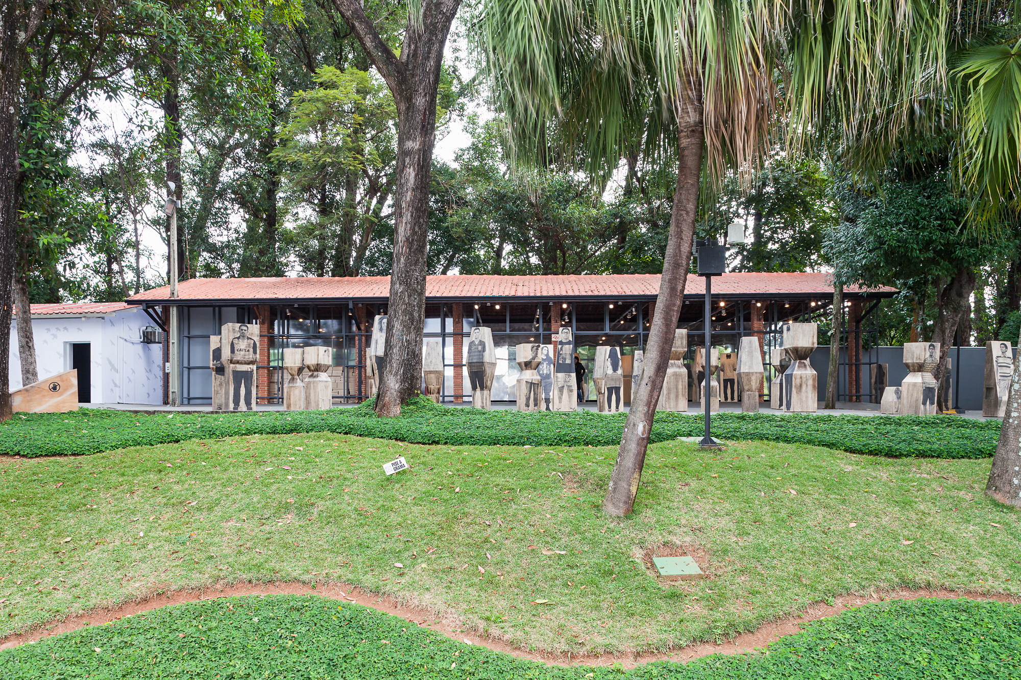 Low pavilion with red tile roof and glazed walls, fronted by rows of printed wooden totems, seen across a lawn under tall trees