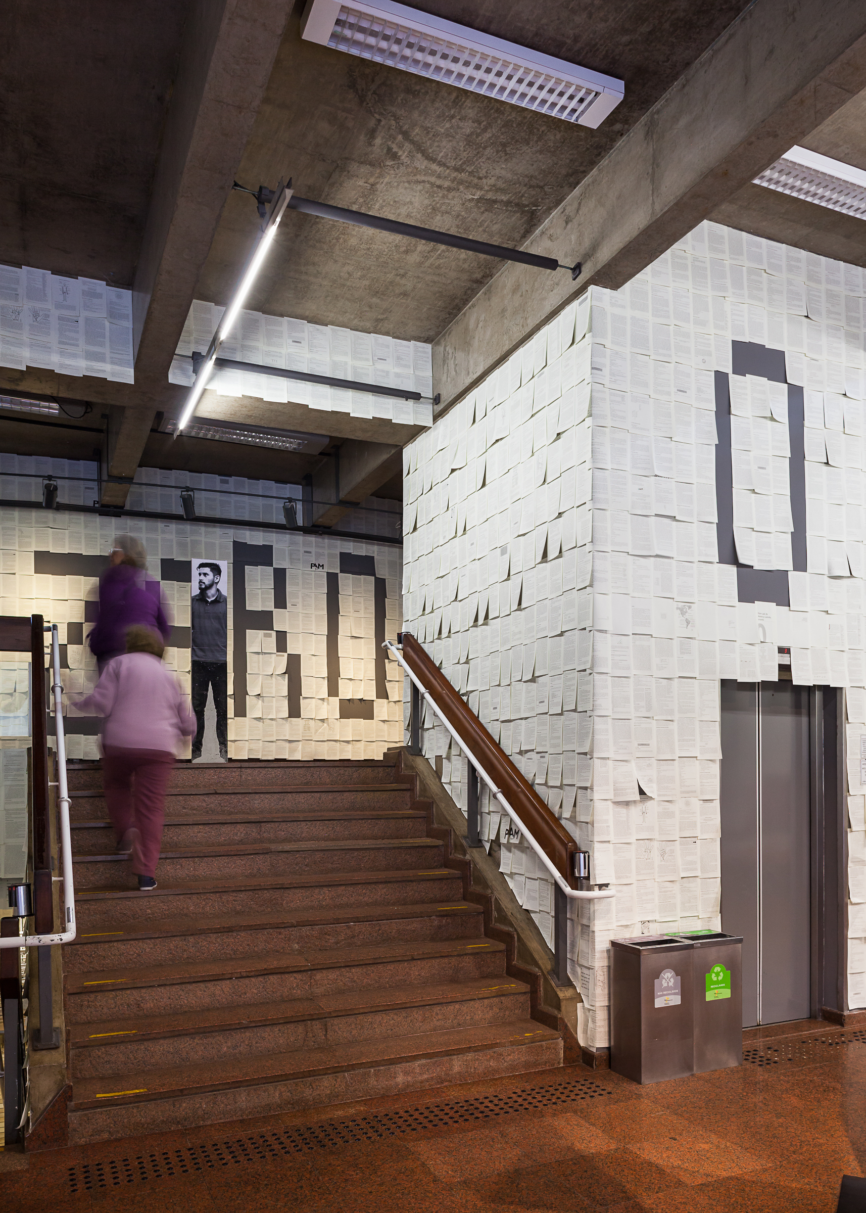 Concrete stairwell with paper-covered walls and a large painted letter, as two visitors in blurred motion climb the granite steps