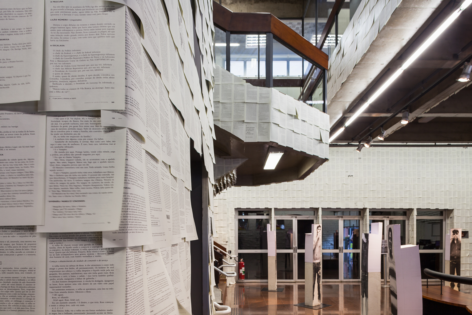 Foreground wall of pasted book pages beside a paper-clad balcony and glazed entrance, with printed figure panels standing in the lobby