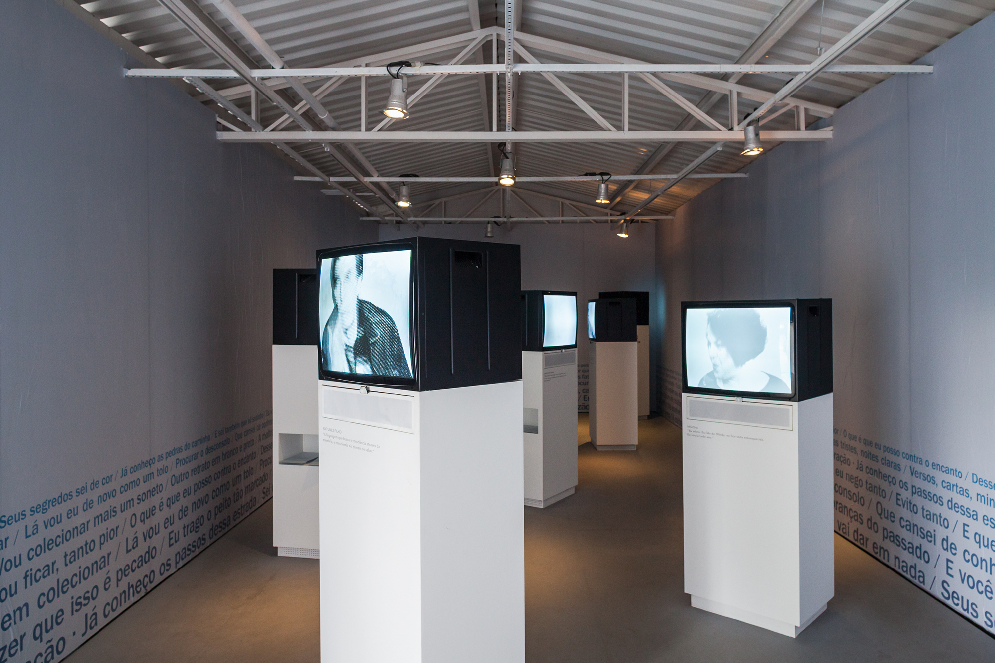 Gallery with steel roof trusses and spotlights, where black-and-white video monitors on white plinths stand between walls printed with blue text
