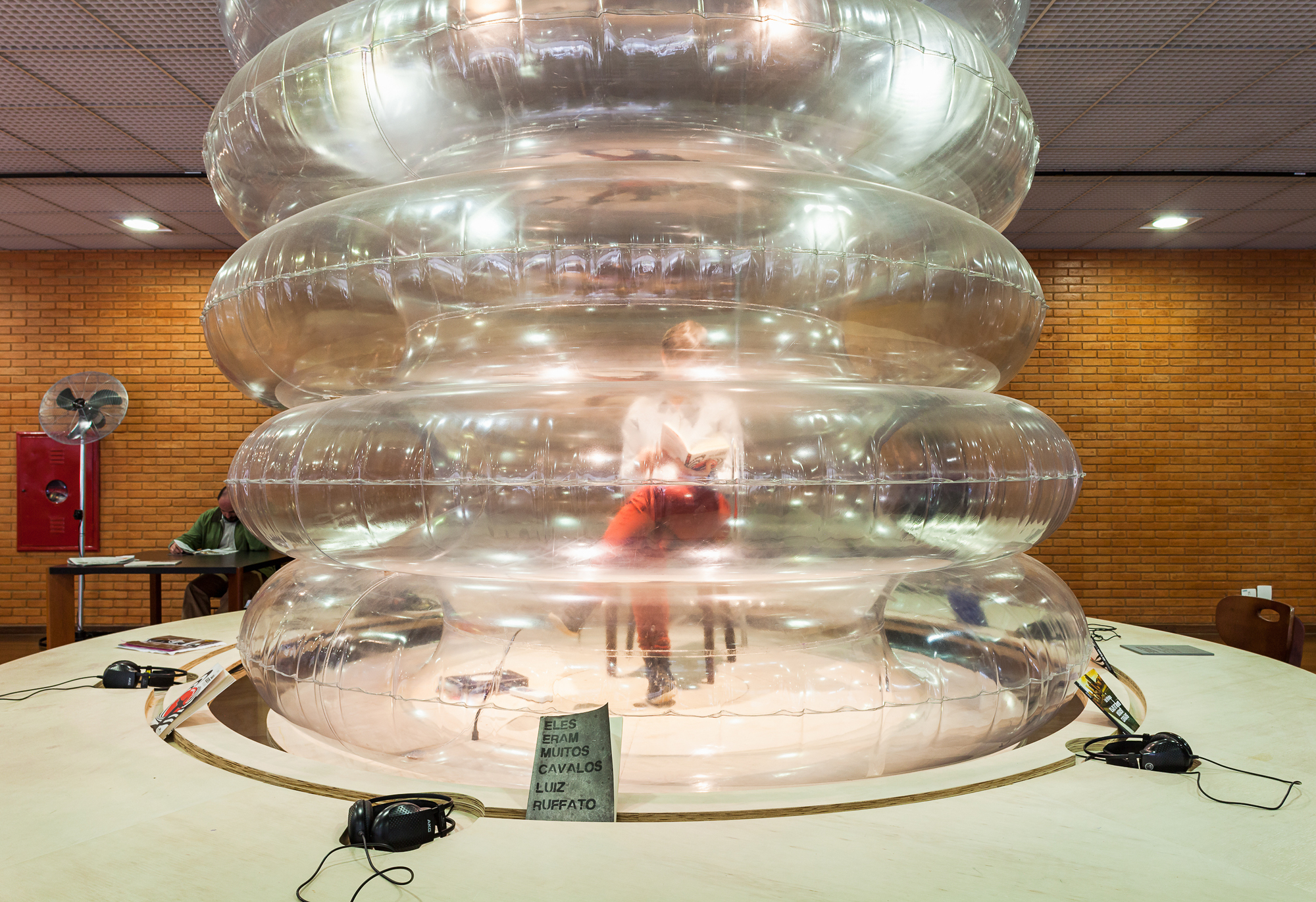 Stack of clear inflatable rings on a round wooden table with headphones, a visitor inside and a reader at a desk against brick