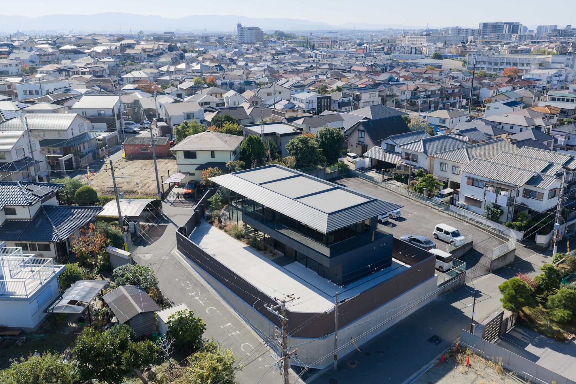 Aerial view of the dark house with a flat-roofed upper volume and walled concrete forecourt among neighbouring tiled rooftops