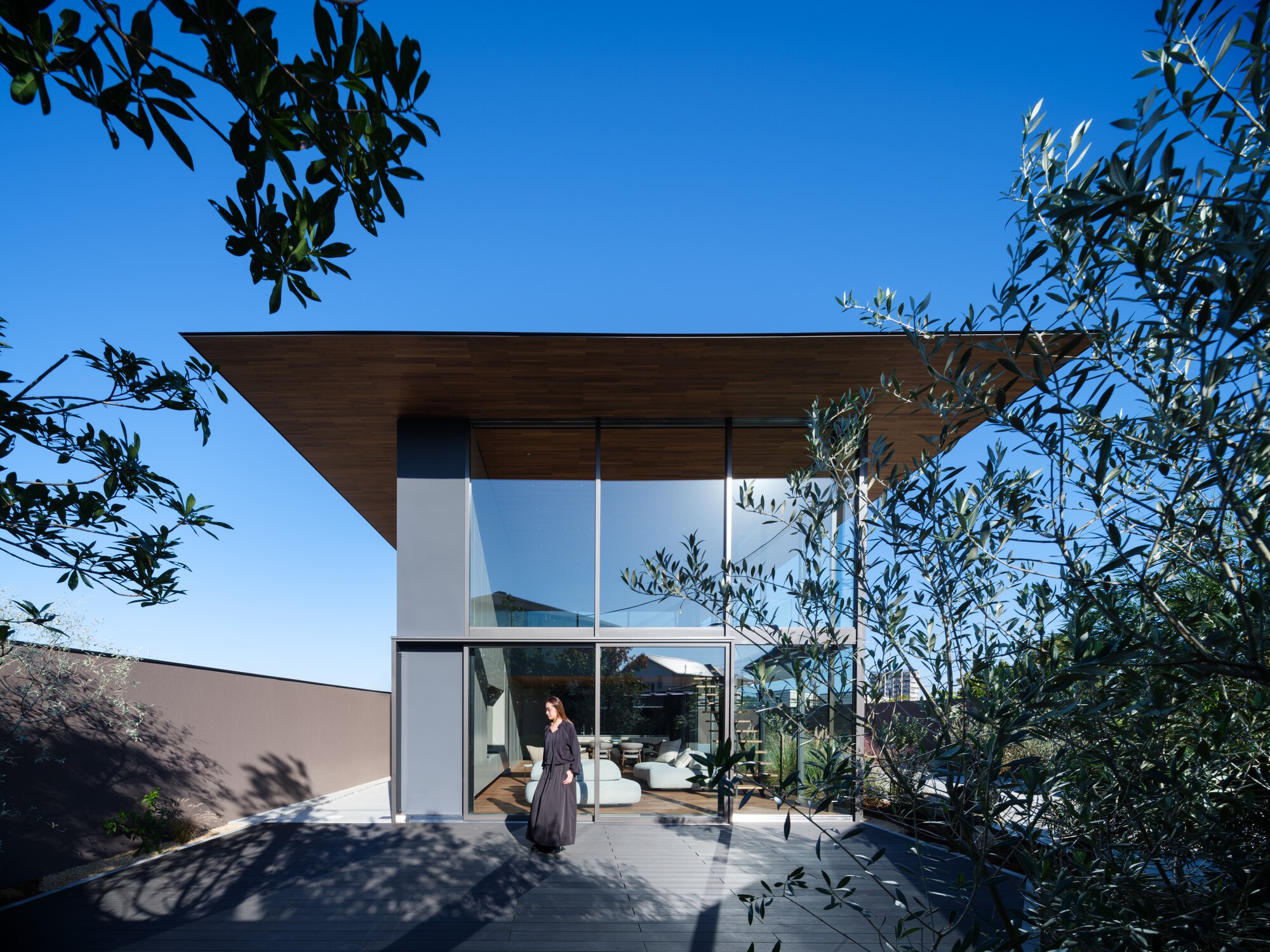 Woman standing before the glazed living space on a grey deck, framed by olive branches and a deep blue sky