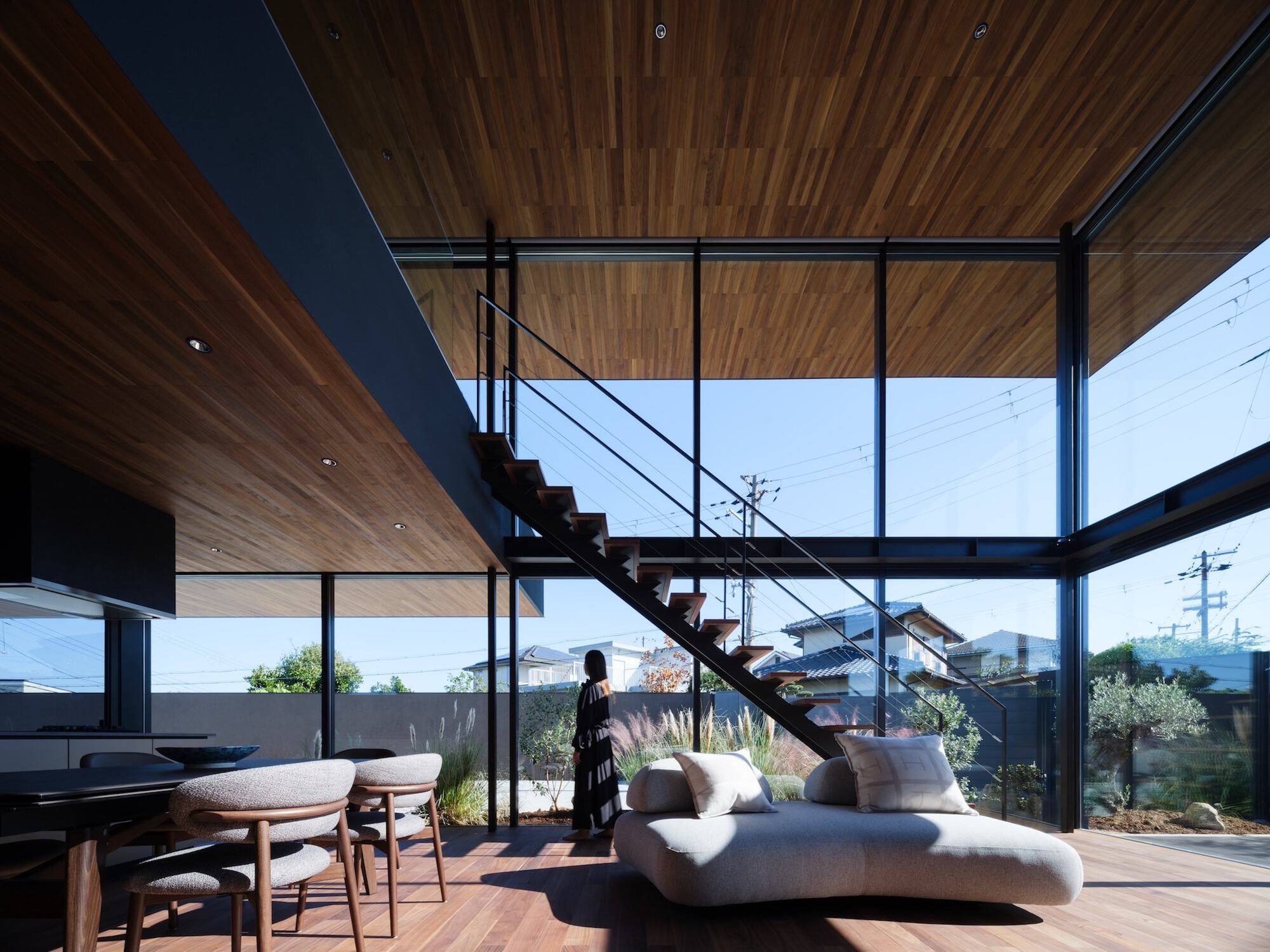 Double-height living room with a steel stair, pebble-shaped sofa and dining chairs, and glass walls opening to the garden in strong sun