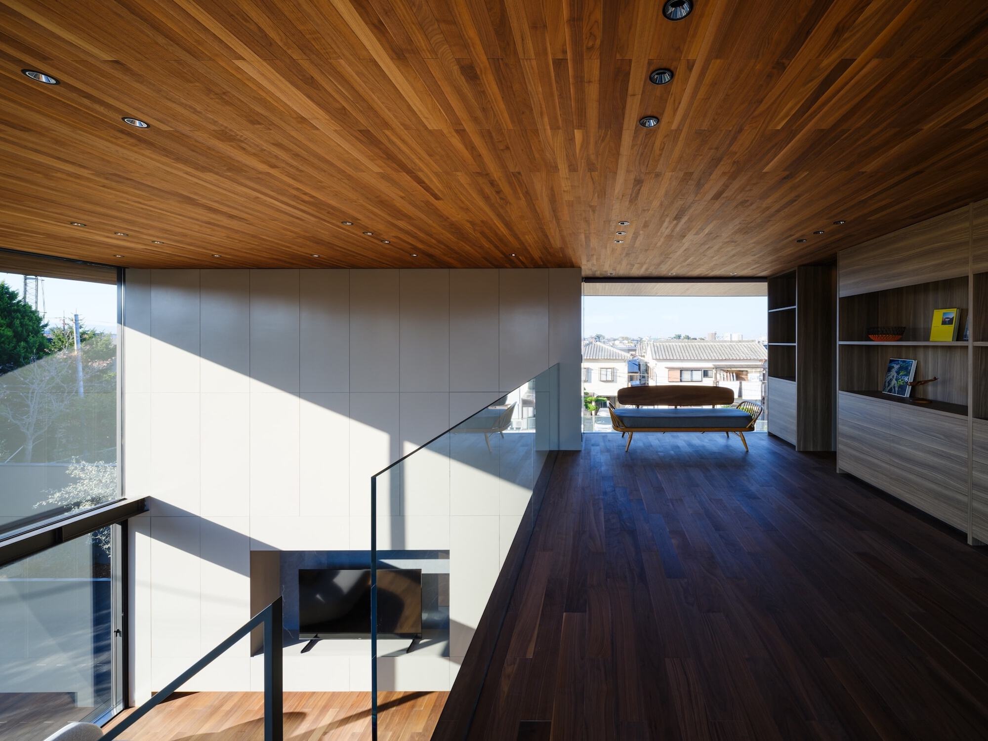 Upper floor lounge with a timber ceiling, glass balustrade, built-in shelving and a daybed facing a window over neighbouring roofs