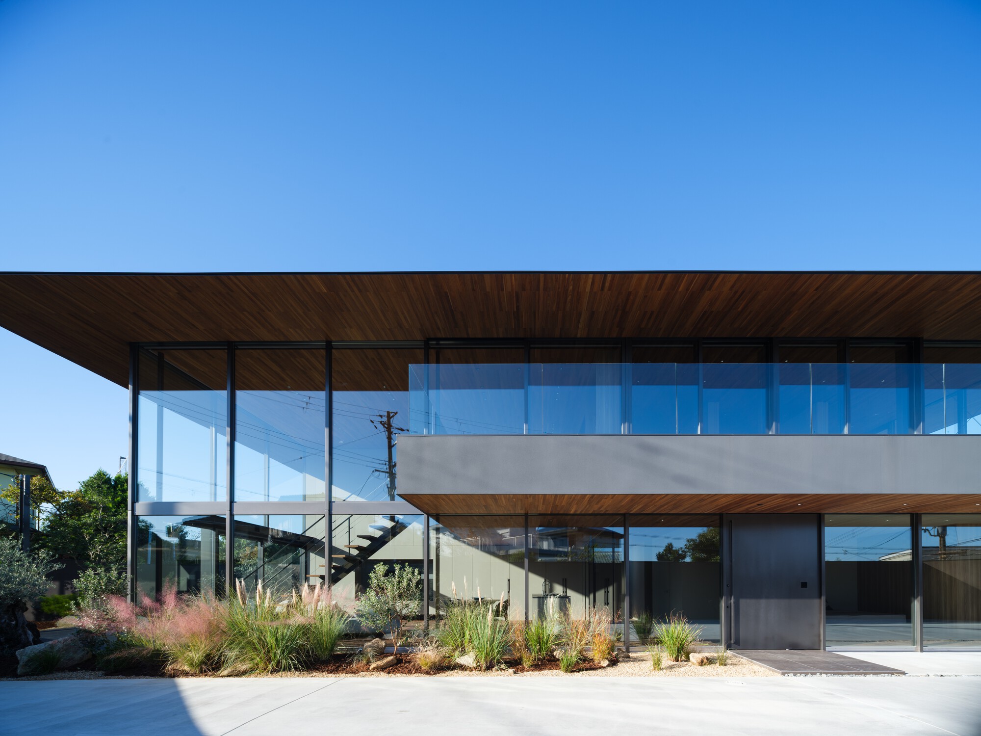 Street-level facade with a deep timber-soffit roof over a glass-walled upper floor, fronted by pink grasses under a clear blue sky