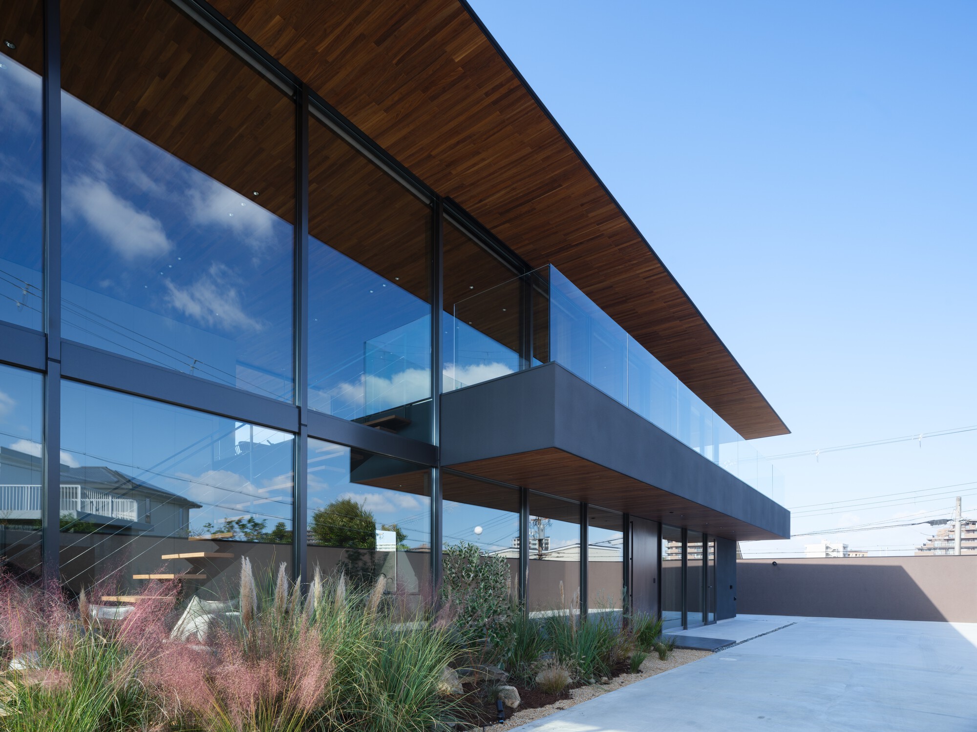 Angled view along the glazed facade and cantilevered grey balcony, with clouds reflected in the glass and grasses in the foreground