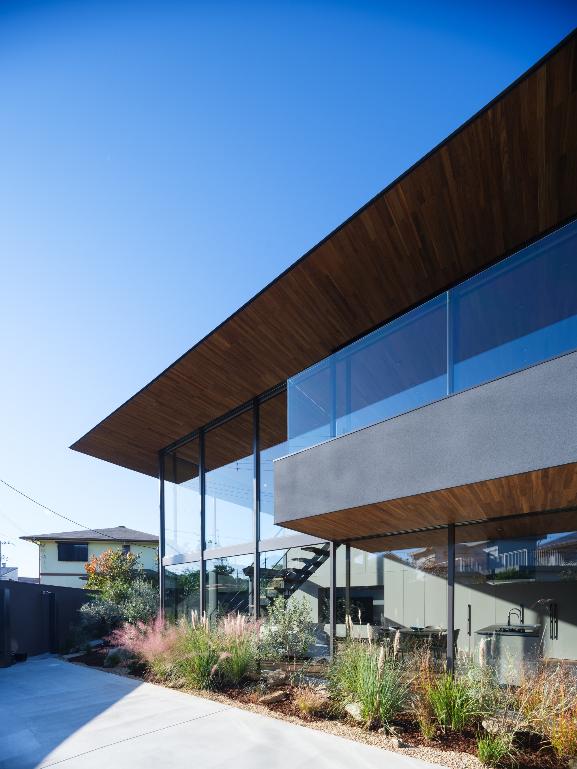 Vertical view of the wide timber roof overhang above a glass balustrade and grey fascia, with ornamental grasses in a gravel bed