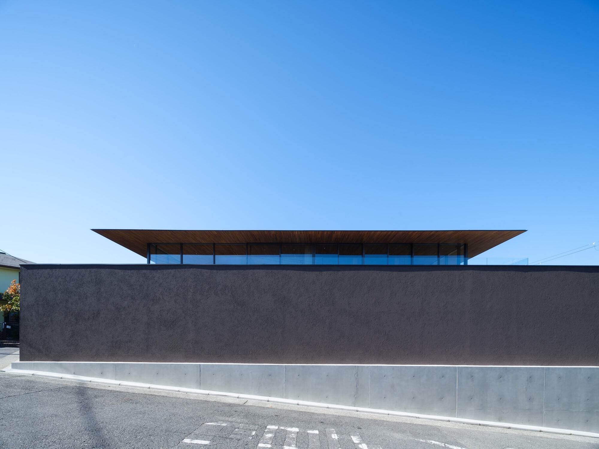 Long dark textured street wall with a concrete base, and a thin timber roof with glazing floating above it in bright sun